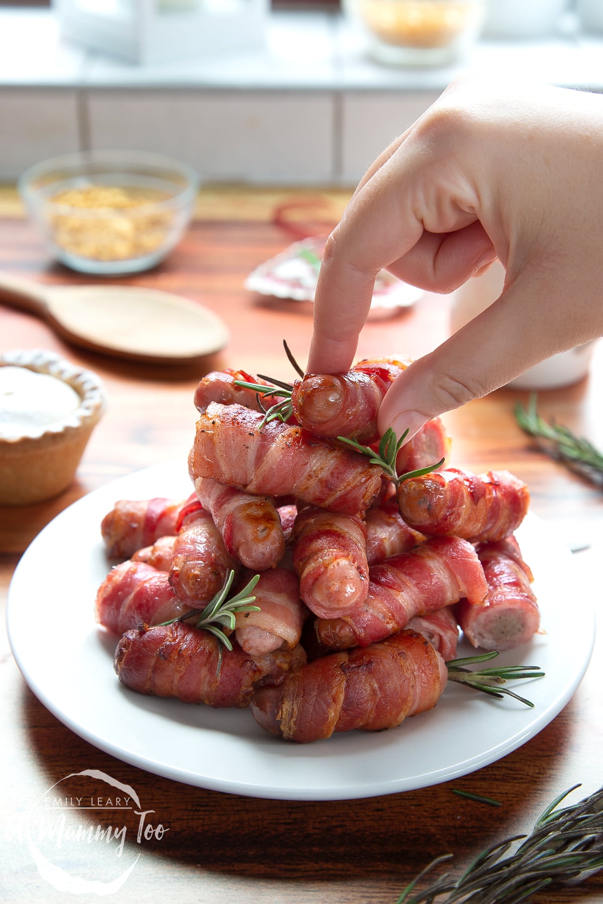 Front angle shot of Bacon wrapped pigs in a blanket with rosemary served on a white plate with a mummy too logo in the lower-left corner