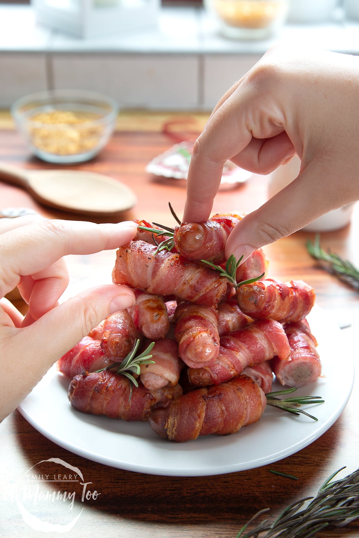 Front angle shot of Bacon wrapped pigs in a blanket with rosemary served on a white plate with a mummy too logo in the lower-left corner