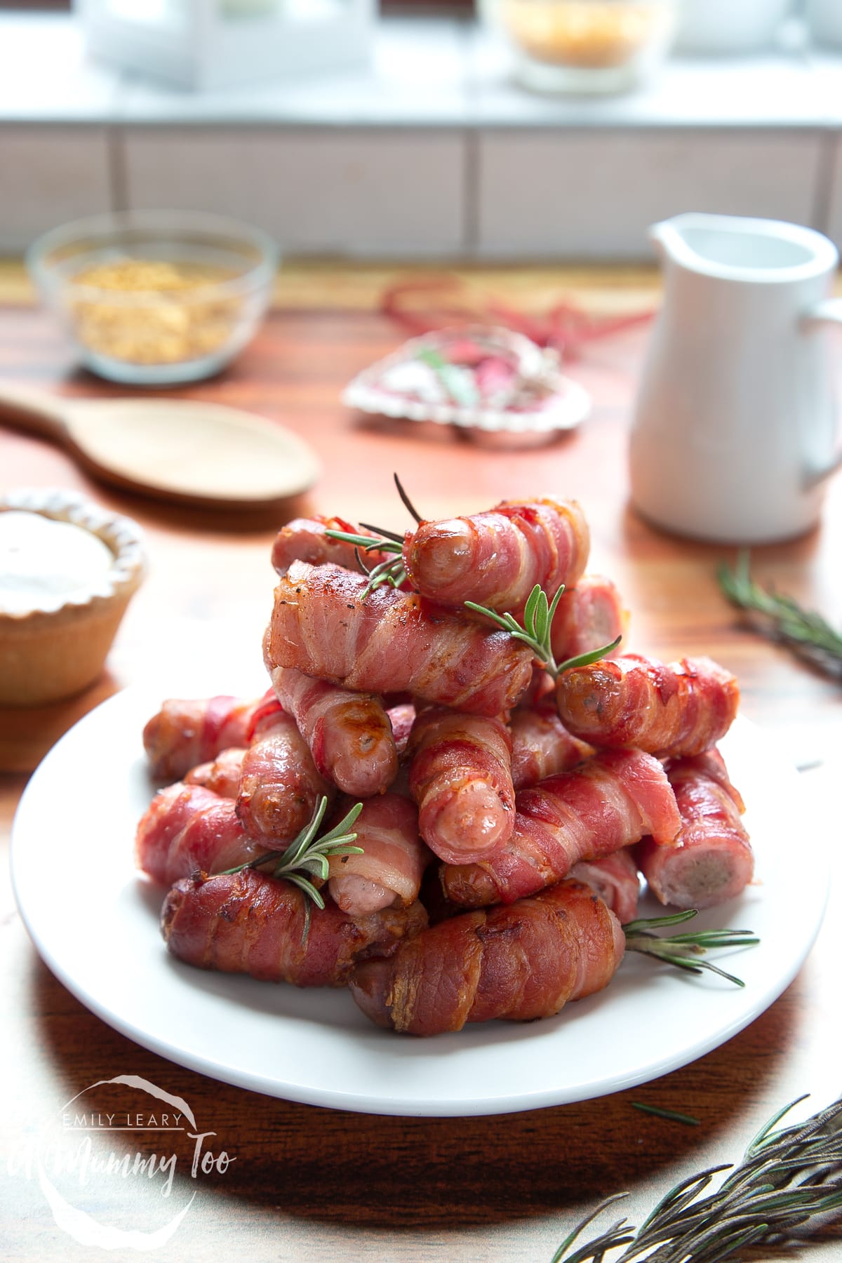 Front angle shot of Bacon with pigs in a blanket with rosemary served on a white plate with a mummy too logo in the lower-left corner