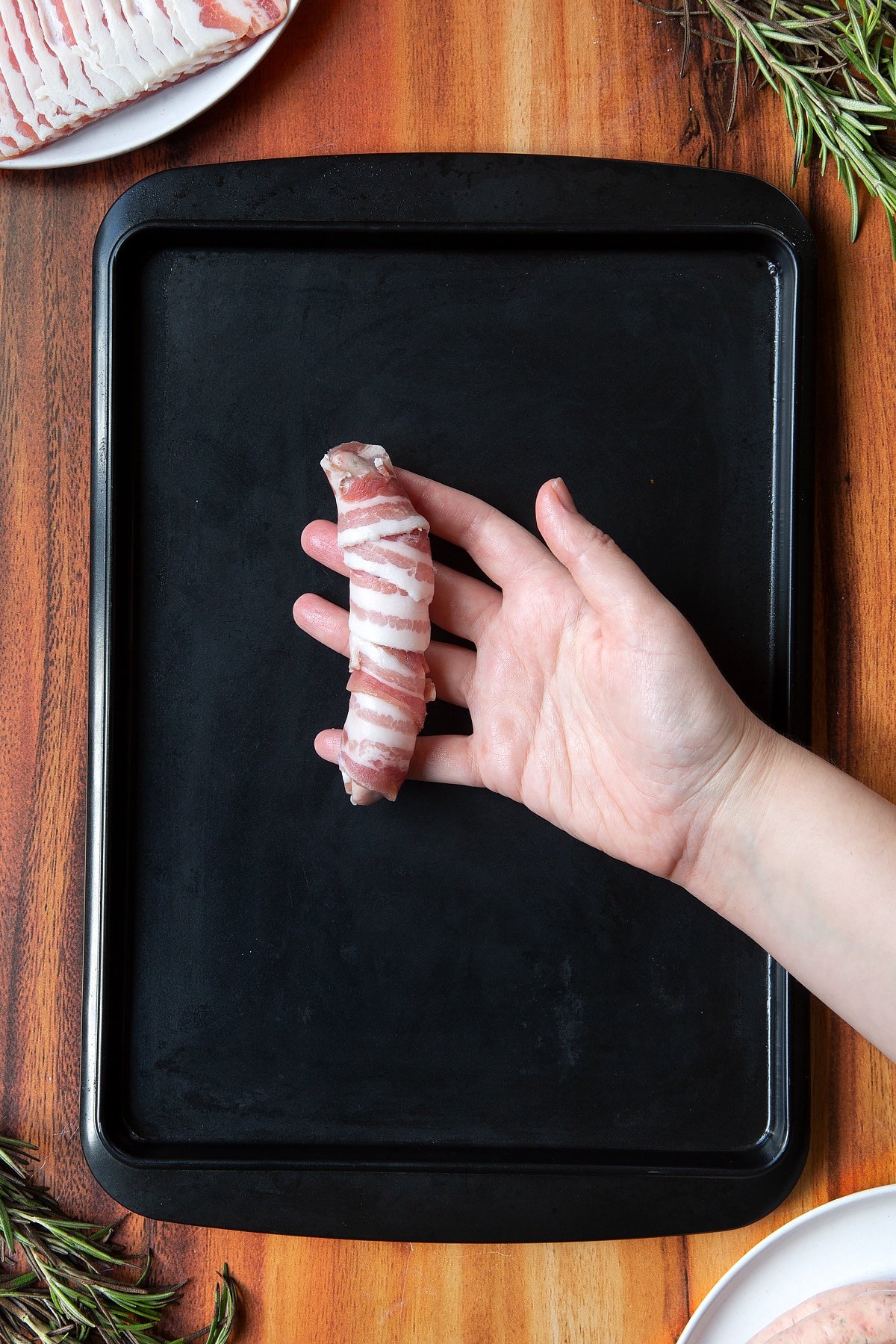 Overhead shot of a hand holding bacon-wrapped sausage above black tray