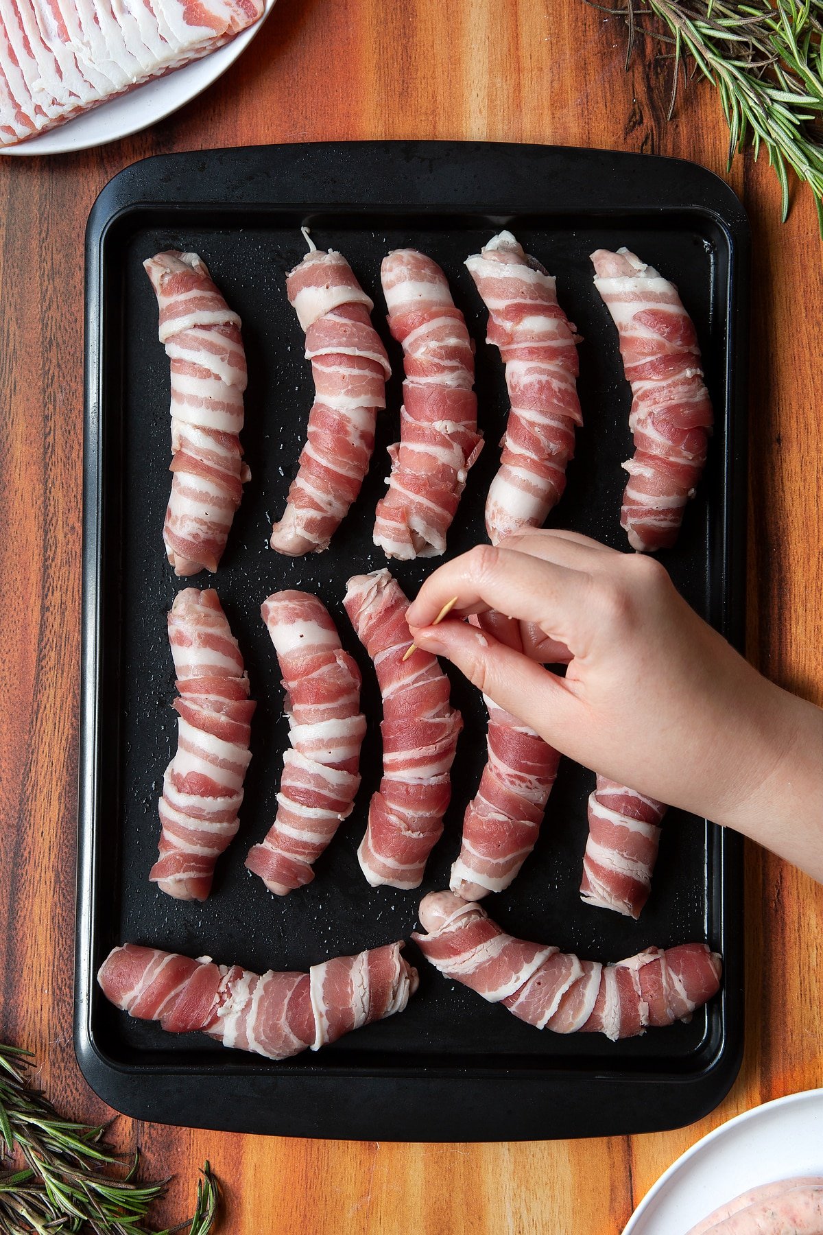 Overhead shot of a hand poking a bacon-wrapped sausage in a black tray