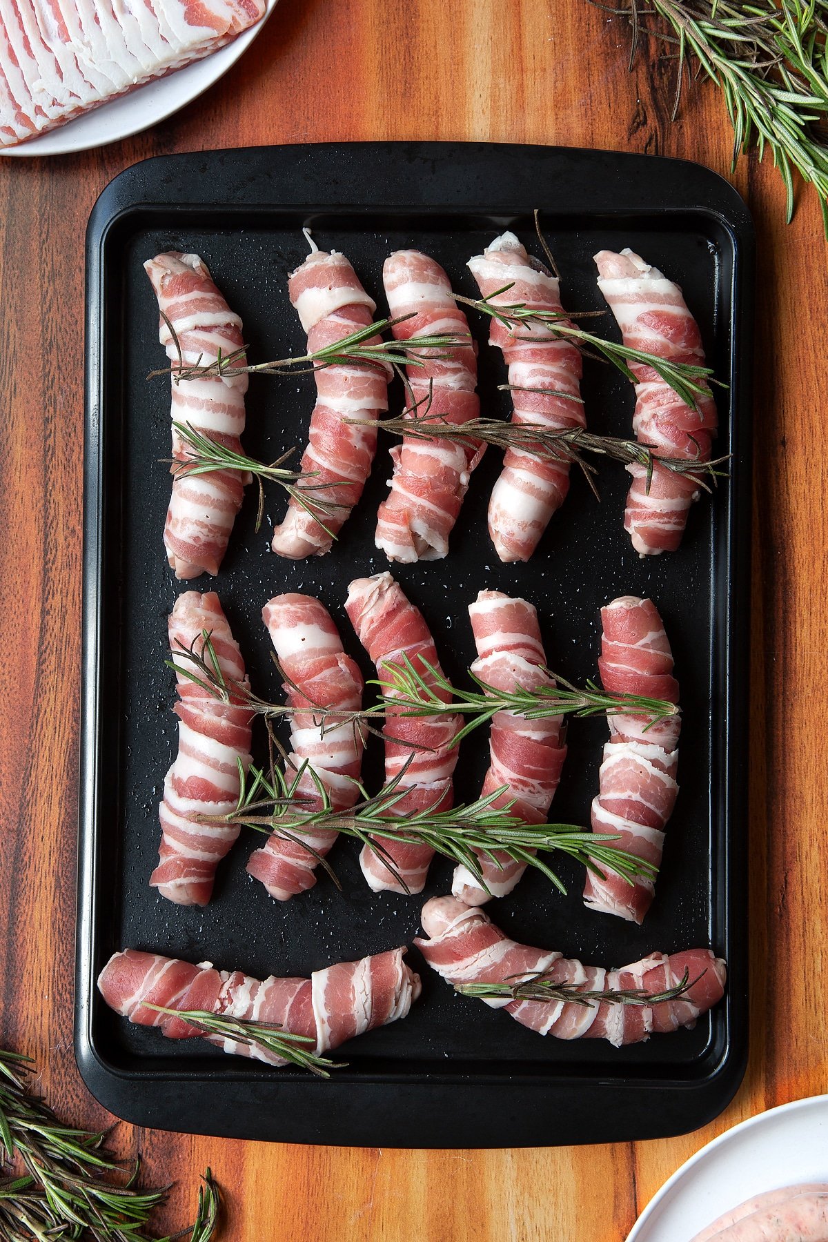 Overhead shot of bacon-wrapped sausages with rosemary on top in a black tray