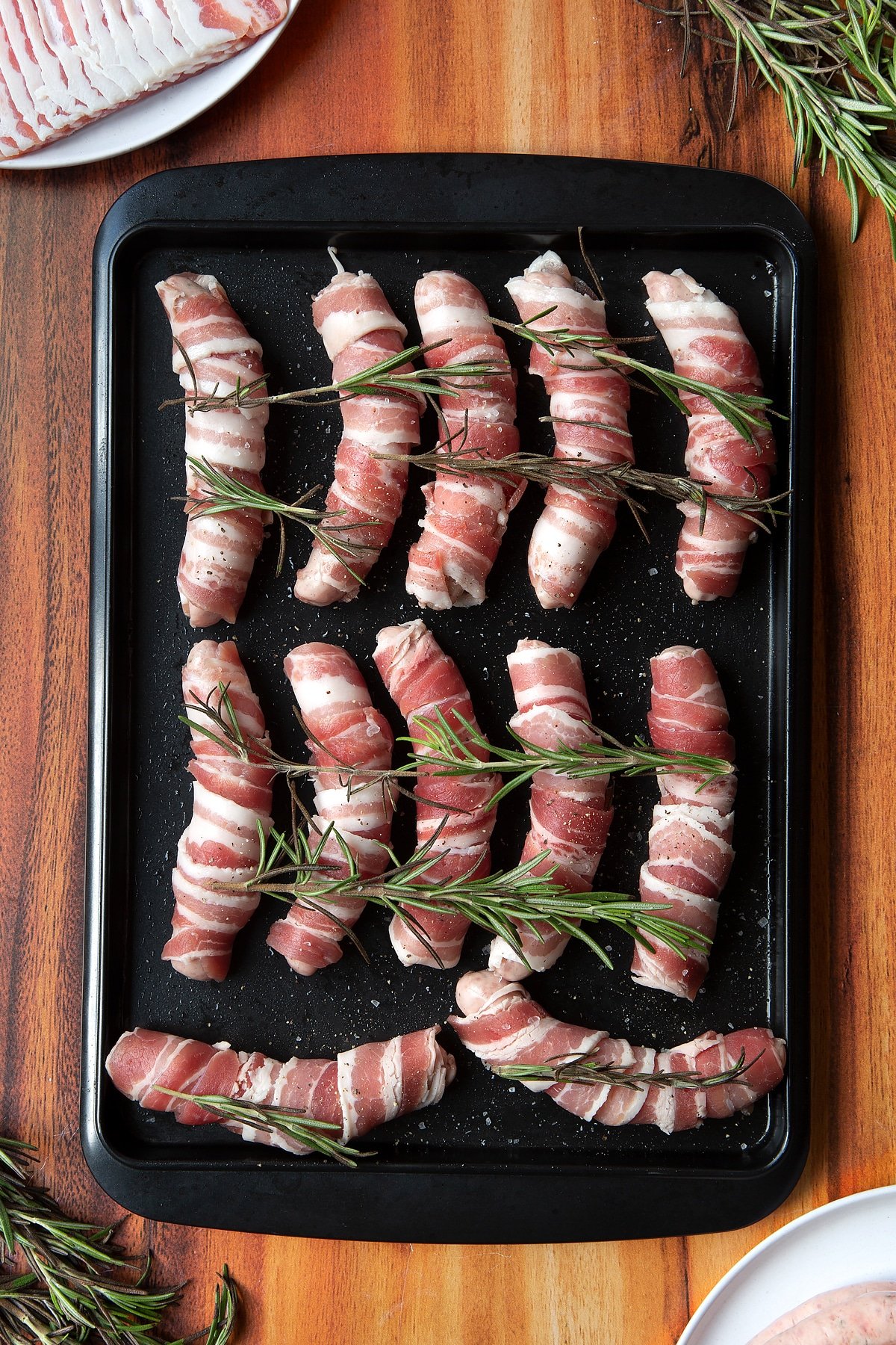 Overhead shot of bacon-wrapped sausages with rosemary on top in a black tray