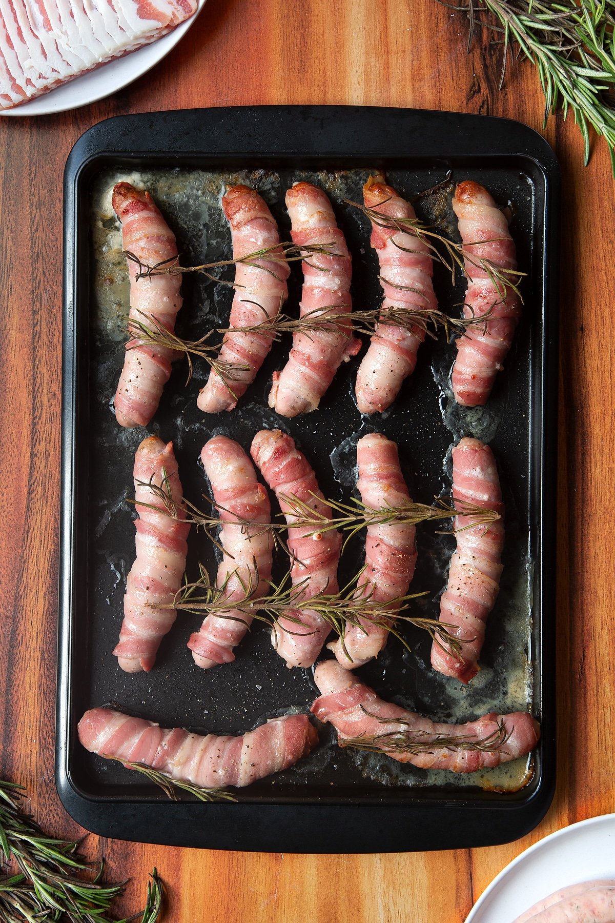 Overhead shot of bacon-wrapped sausages with rosemary on top in a black tray