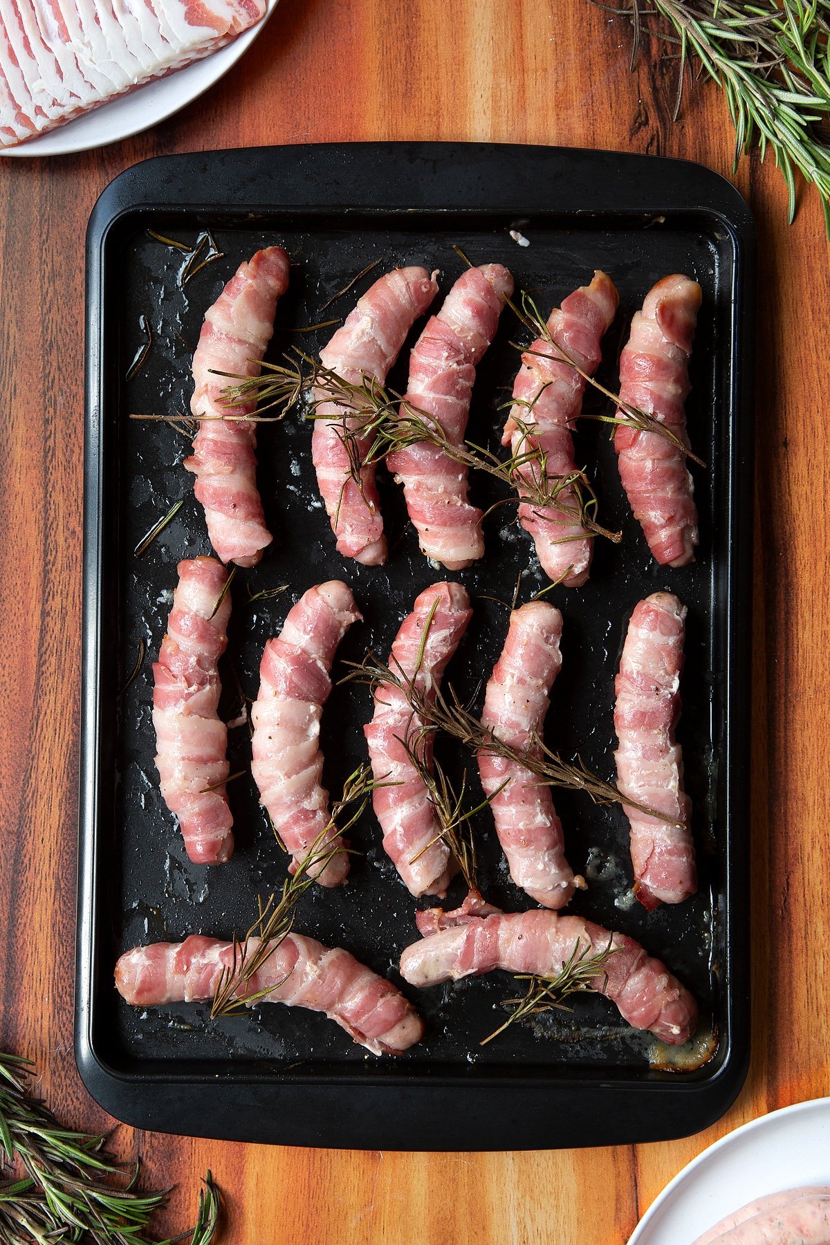 Overhead shot of bacon-wrapped sausages with rosemary on top in a black tray