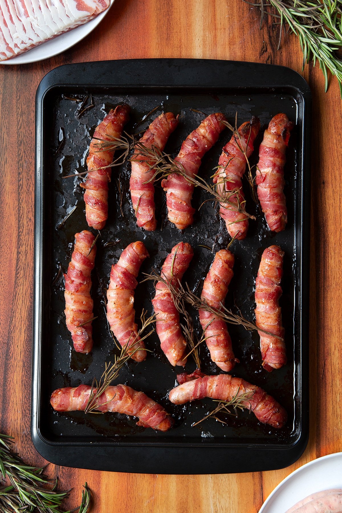 Overhead shot of bacon-wrapped cooked sausages with rosemary on top in a black tray. The bacon is crispy.