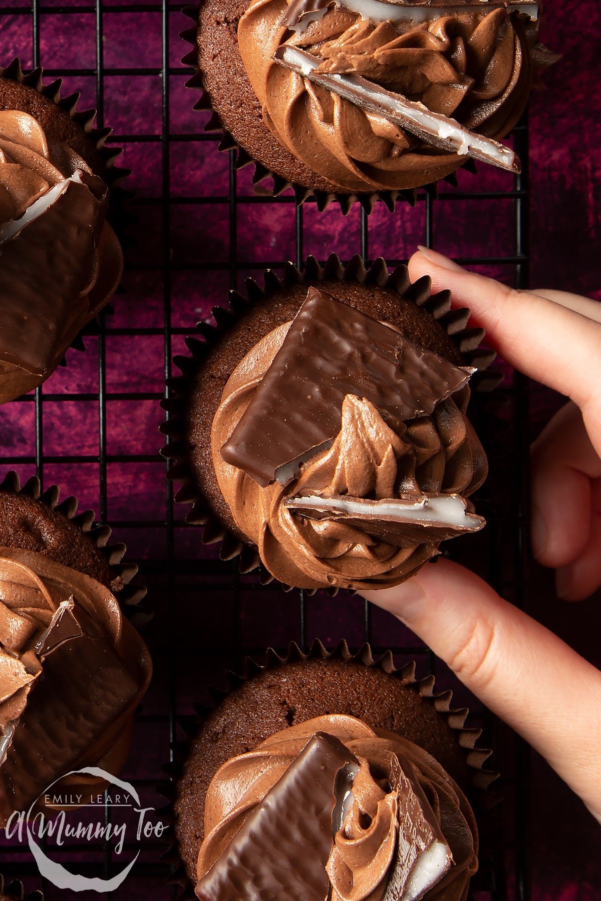 An After Eight cupcake on a cooling rack on purple surface. More cupcakes surround it. A hand reaches for one.
