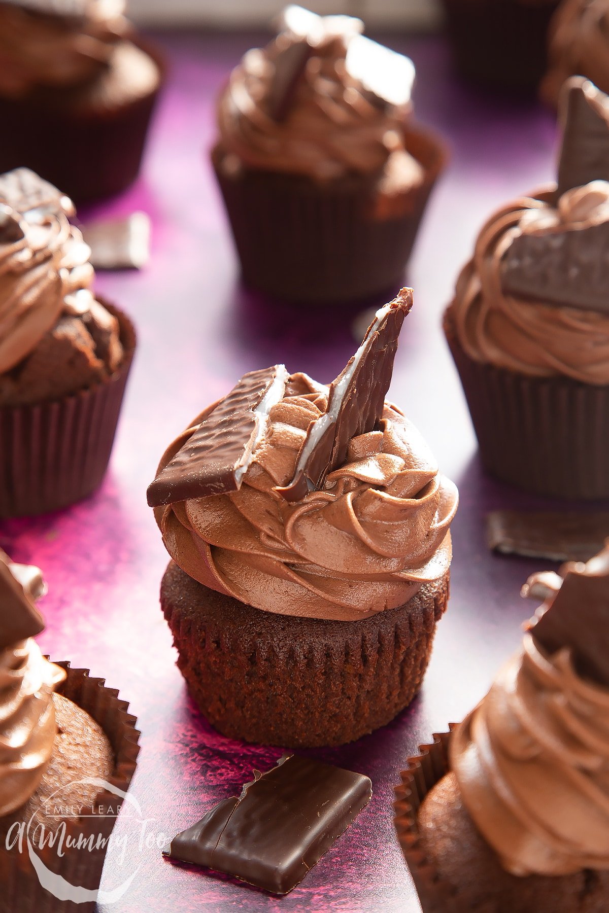 An unwrapped After Eight cupcake on a purple surface. More cupcakes surround it.