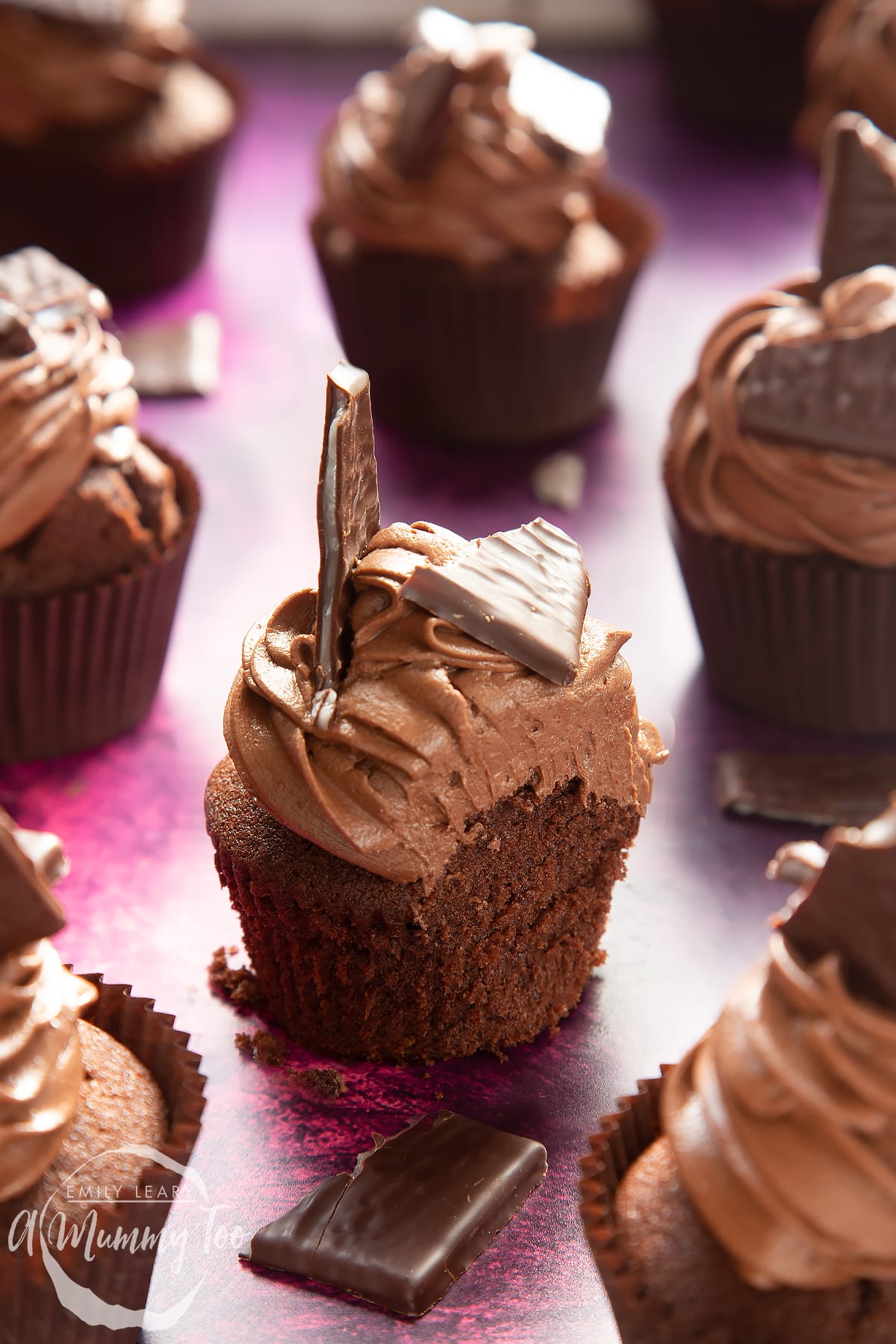 An After Eight cupcake on a purple surface. The cupcake has been cut into showing the sponge inside. More cupcakes surround it.