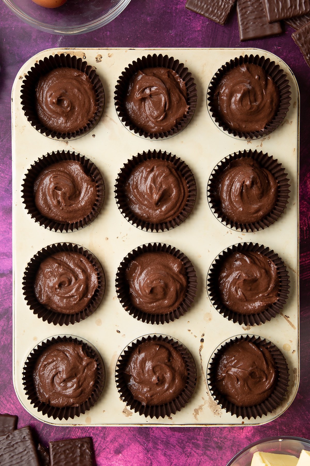 A 12 hole muffin tray lined with paper cases filled with chocolate cake batter. Ingredients to make After Eight cupcakes surround the tray.