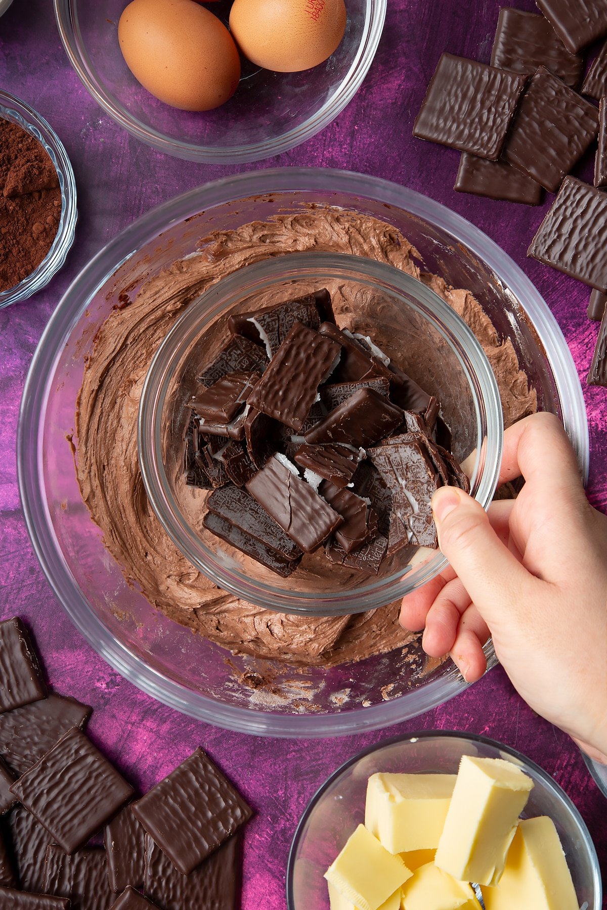 Hand holding of small bowl of broken After Eights above a bowl of chocolate buttercream. Ingredients to make After Eight cupcakes surround the bowl.