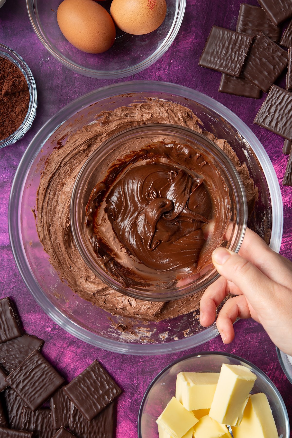 Hand holding of small bowl of melted After Eights above a bowl of chocolate buttercream. Ingredients to make After Eight cupcakes surround the bowl.