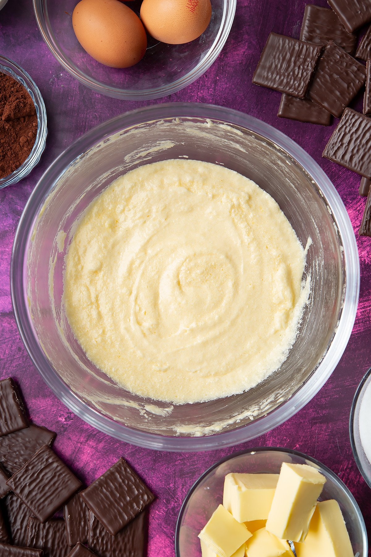 Sugar, butter and eggs beaten together in a glass mixing bowl. Ingredients to make After Eight cupcakes surround the bowl.