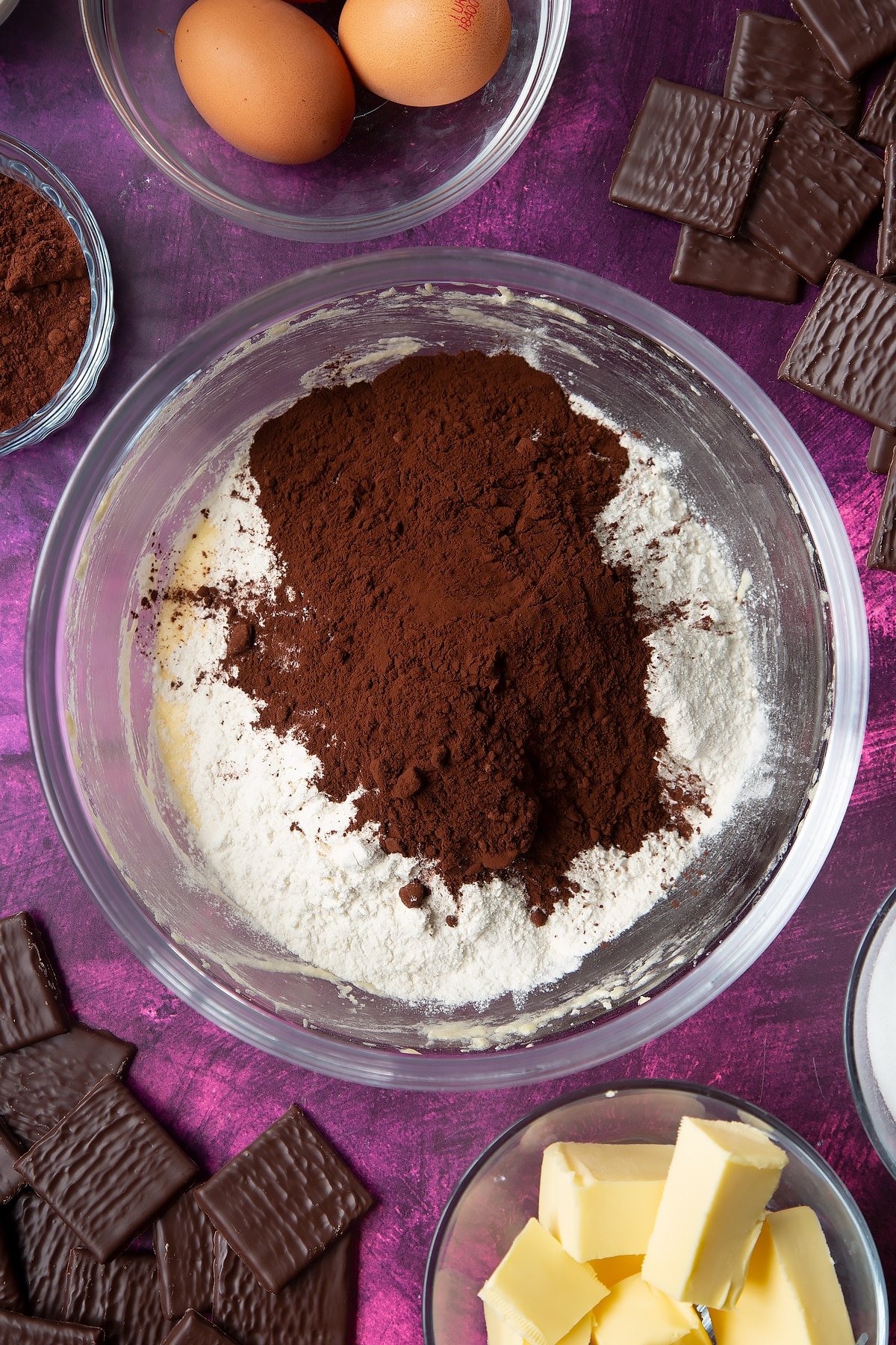 Sugar, butter and eggs beaten together in a glass mixing bowl with flour and cocoa on top. Ingredients to make After Eight cupcakes surround the bowl.