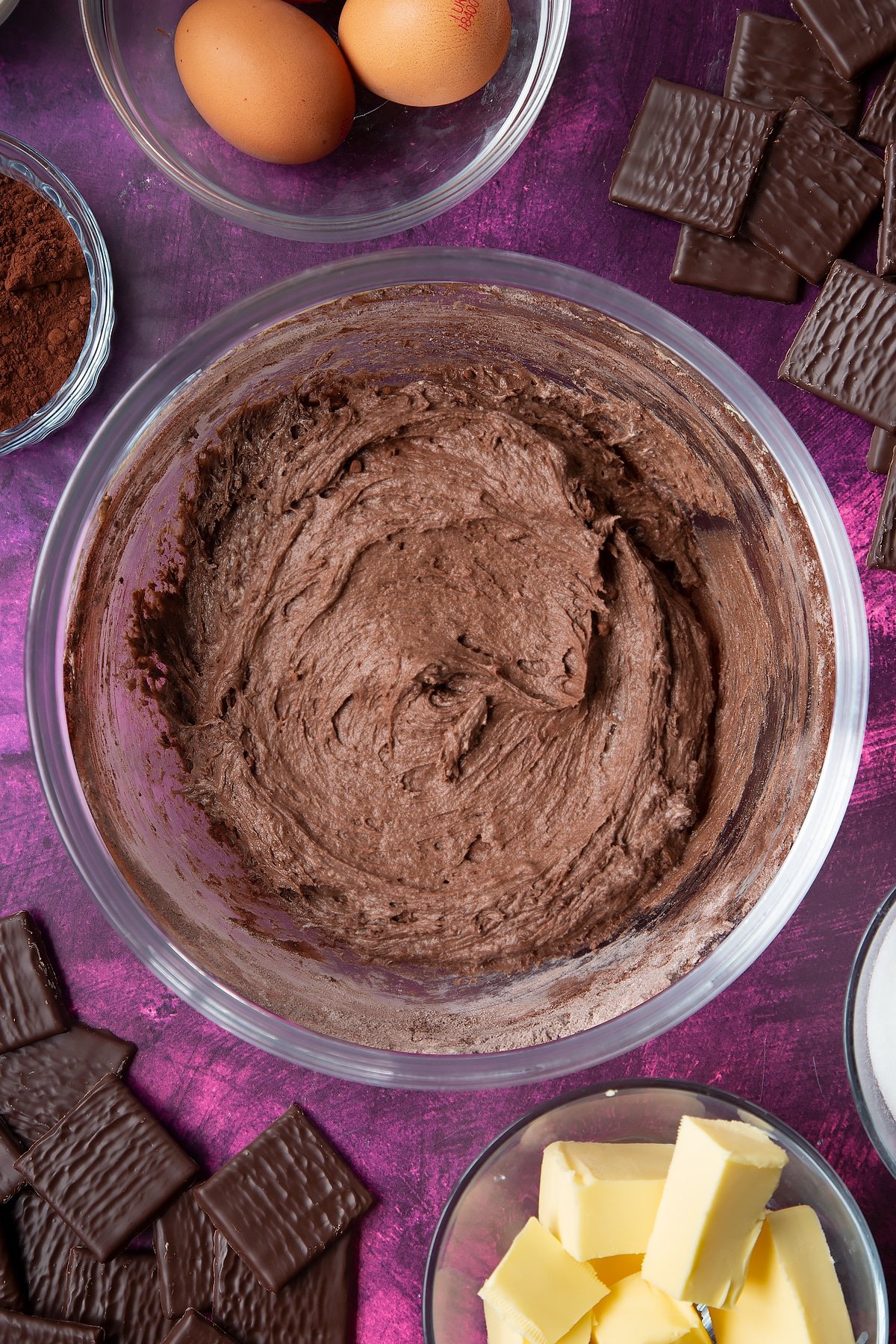 Sugar, butter, eggs, flour and cocoa mixed together in a glass mixing bowl. Ingredients to make After Eight cupcakes surround the bowl.