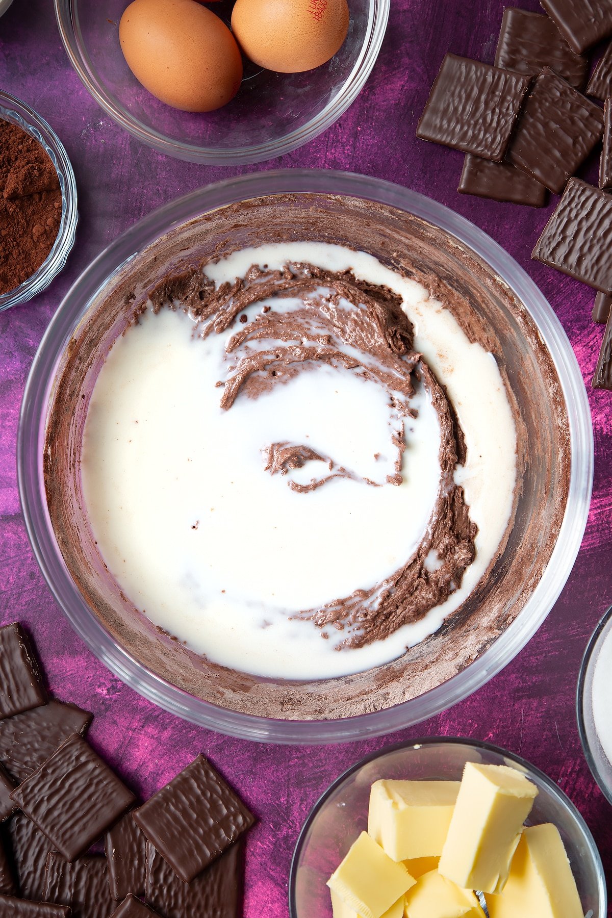 Sugar, butter, eggs, flour and cocoa mixed together in a glass mixing bowl with milk on top. Ingredients to make After Eight cupcakes surround the bowl.