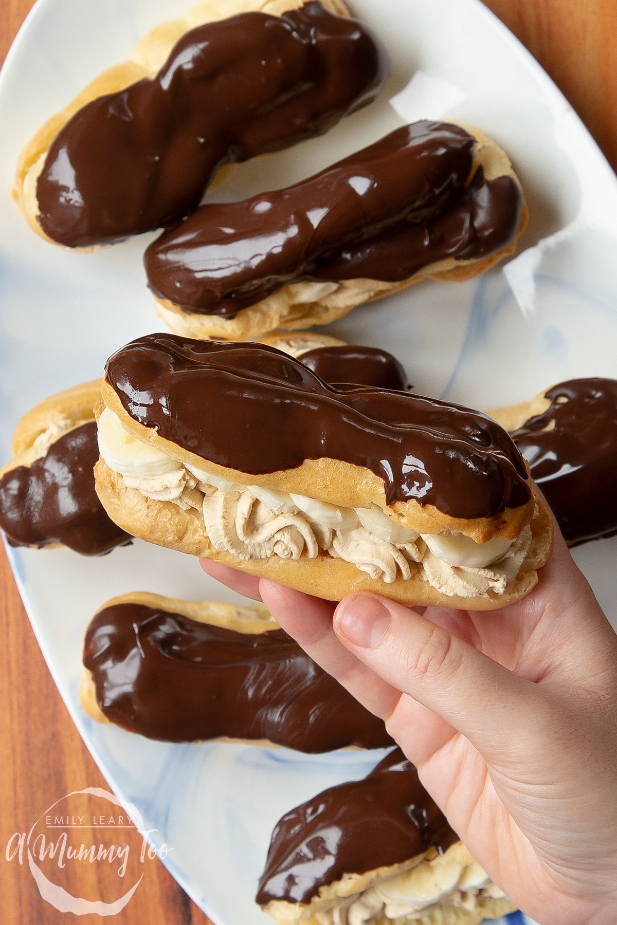 Overhead shot of a hand holding a banoffee éclair with a mummy too logo in the lower-left corner