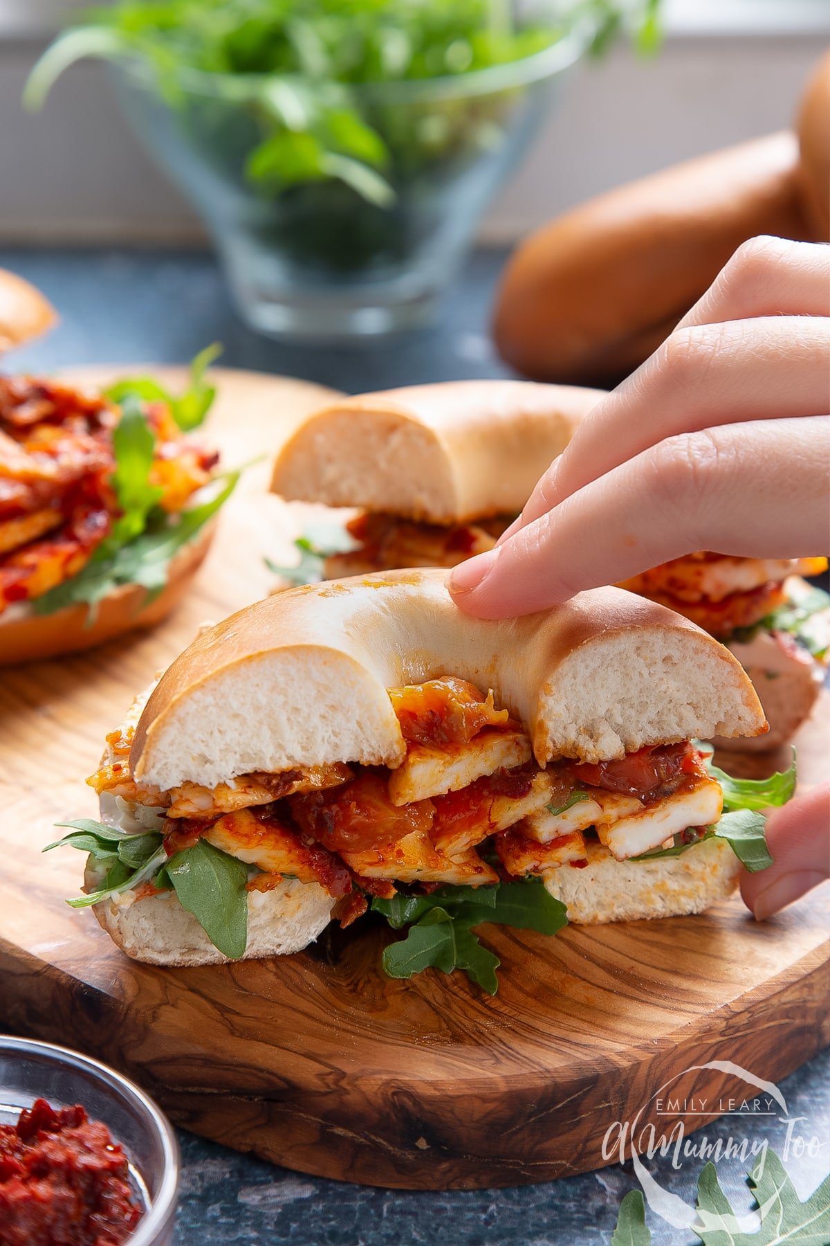 Two halloumi bagels on a wooden board. One is cut in half. A hand reaches for it.