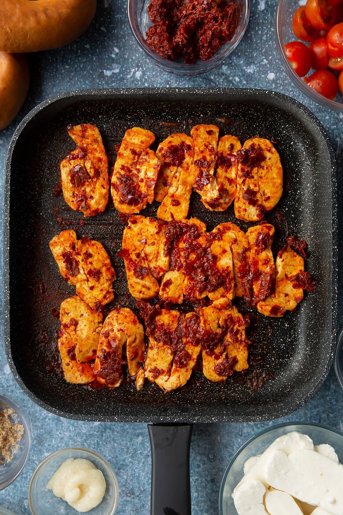 Slices of halloumi brushed with harissa paste and fried in a griddle pan. Ingredients to make halloumi bagels surround the pan.