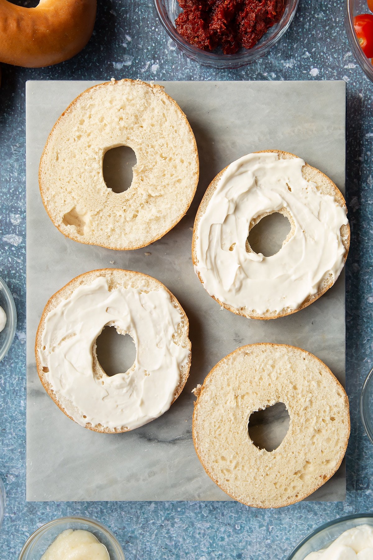 Sliced open bagels on a marble board. Half are spread with mayo. Ingredients to make halloumi bagels surround the board.