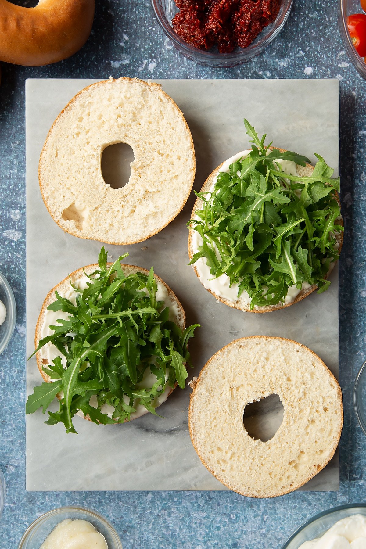 Sliced open bagels on a marble board. Half are spread with mayo and topped with rocket. Ingredients to make halloumi bagels surround the board.