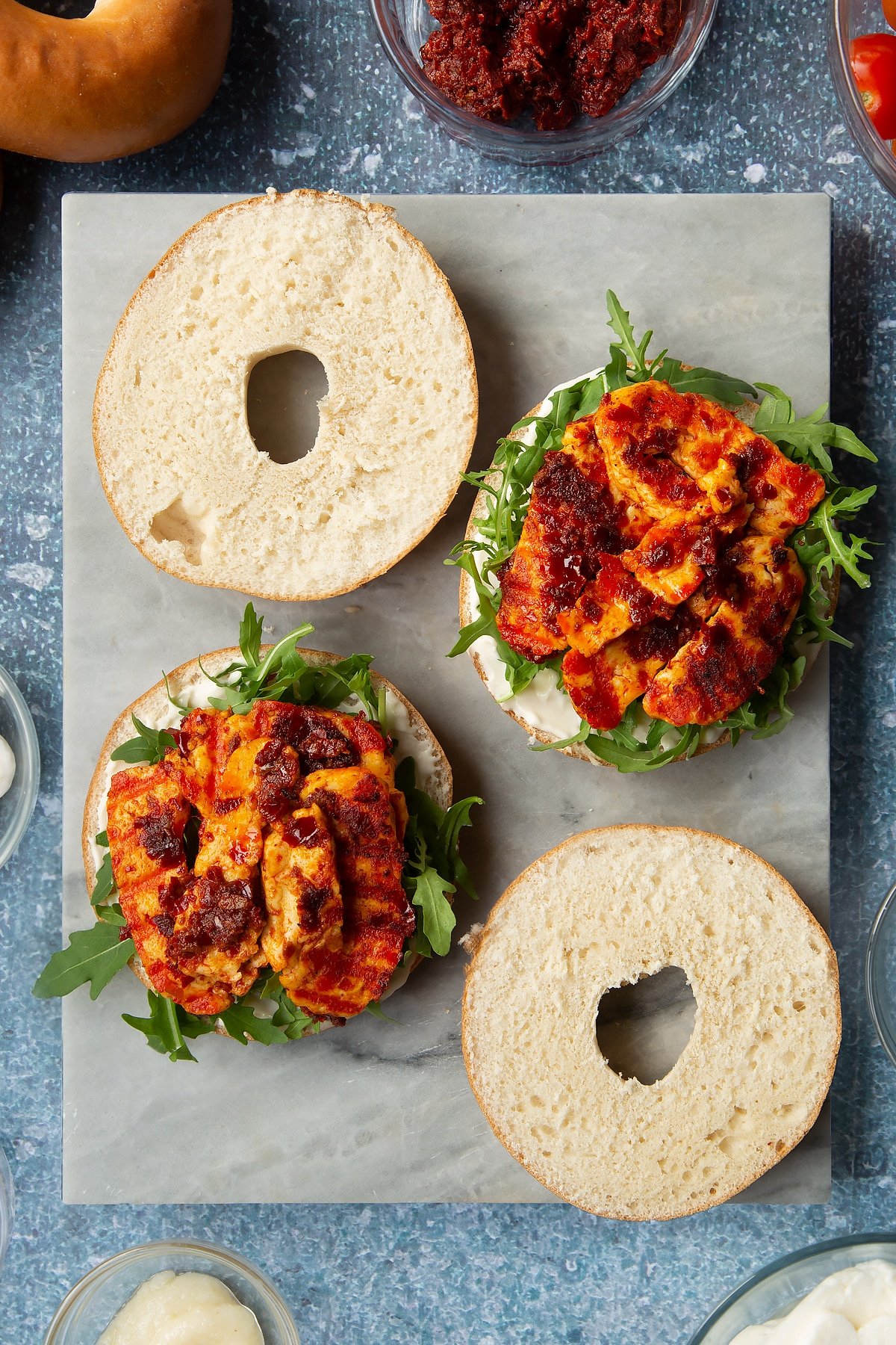 Sliced open bagels on a marble board. Half are spread with mayo and topped with rocket and harissa halloumi. Ingredients to make halloumi bagels surround the board.