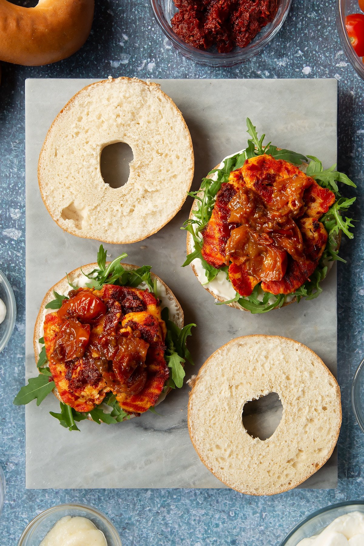 Sliced open bagels on a marble board. Half are spread with mayo and topped with rocket, harissa halloumi and cherry tomato salsa. Ingredients to make halloumi bagels surround the board.