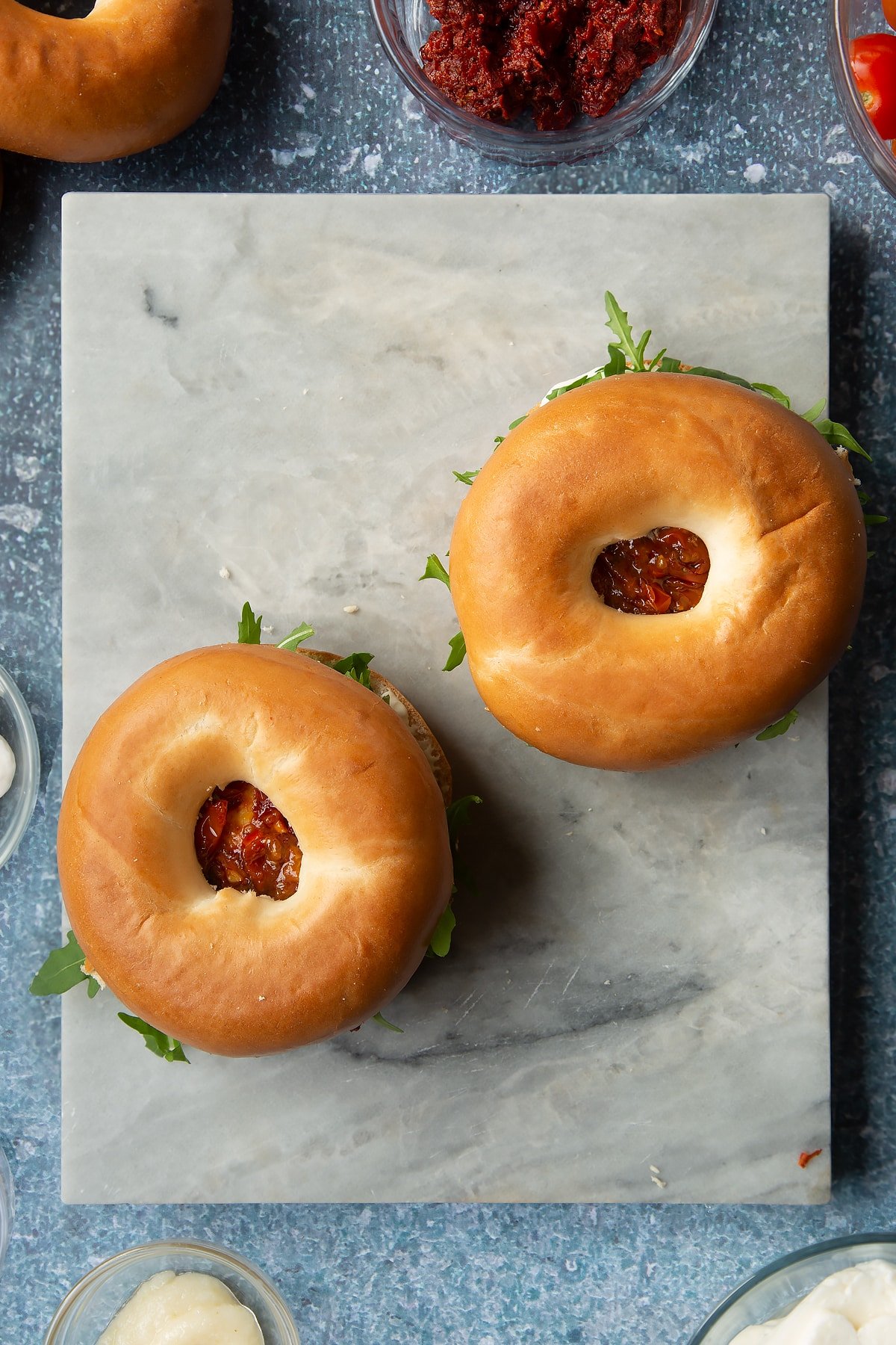 Two bagels filled with mayo and topped with rocket, harissa halloumi and cherry tomato salsa. Ingredients to make halloumi bagels surround the board.