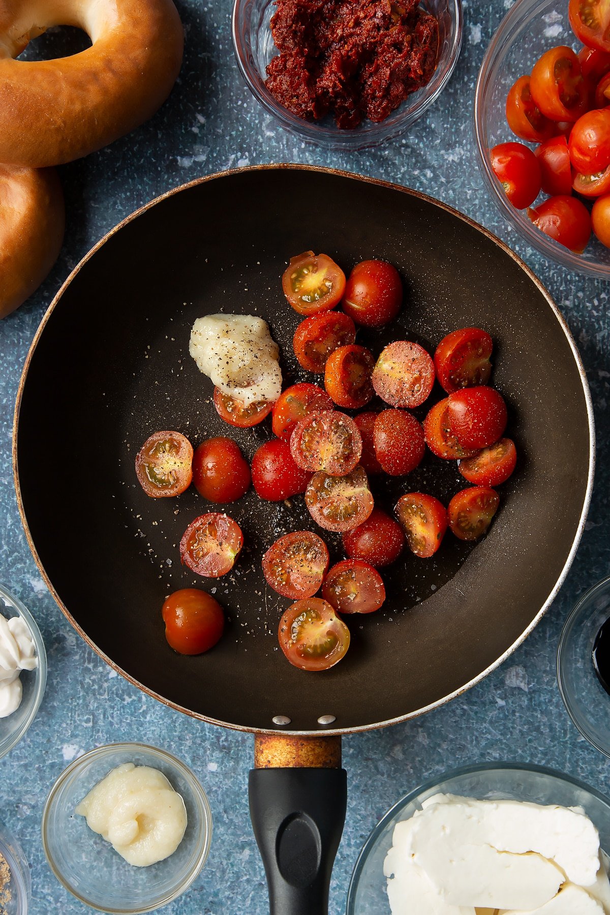 Oil, cherry tomatoes, garlic and pepper in a frying pan. Ingredients to make halloumi bagels surround the pan.