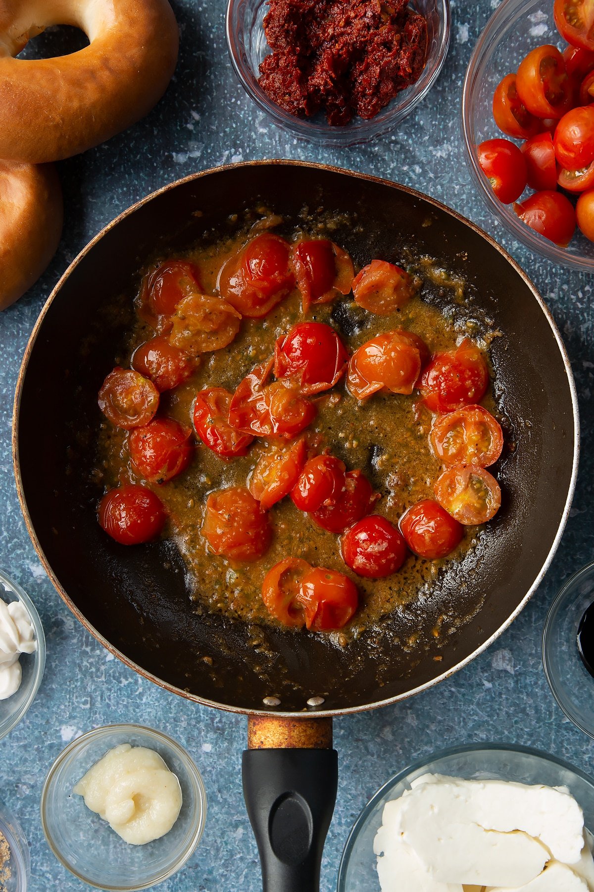 Fried down cherry tomatoes, garlic and pepper in a frying pan. Ingredients to make halloumi bagels surround the pan.