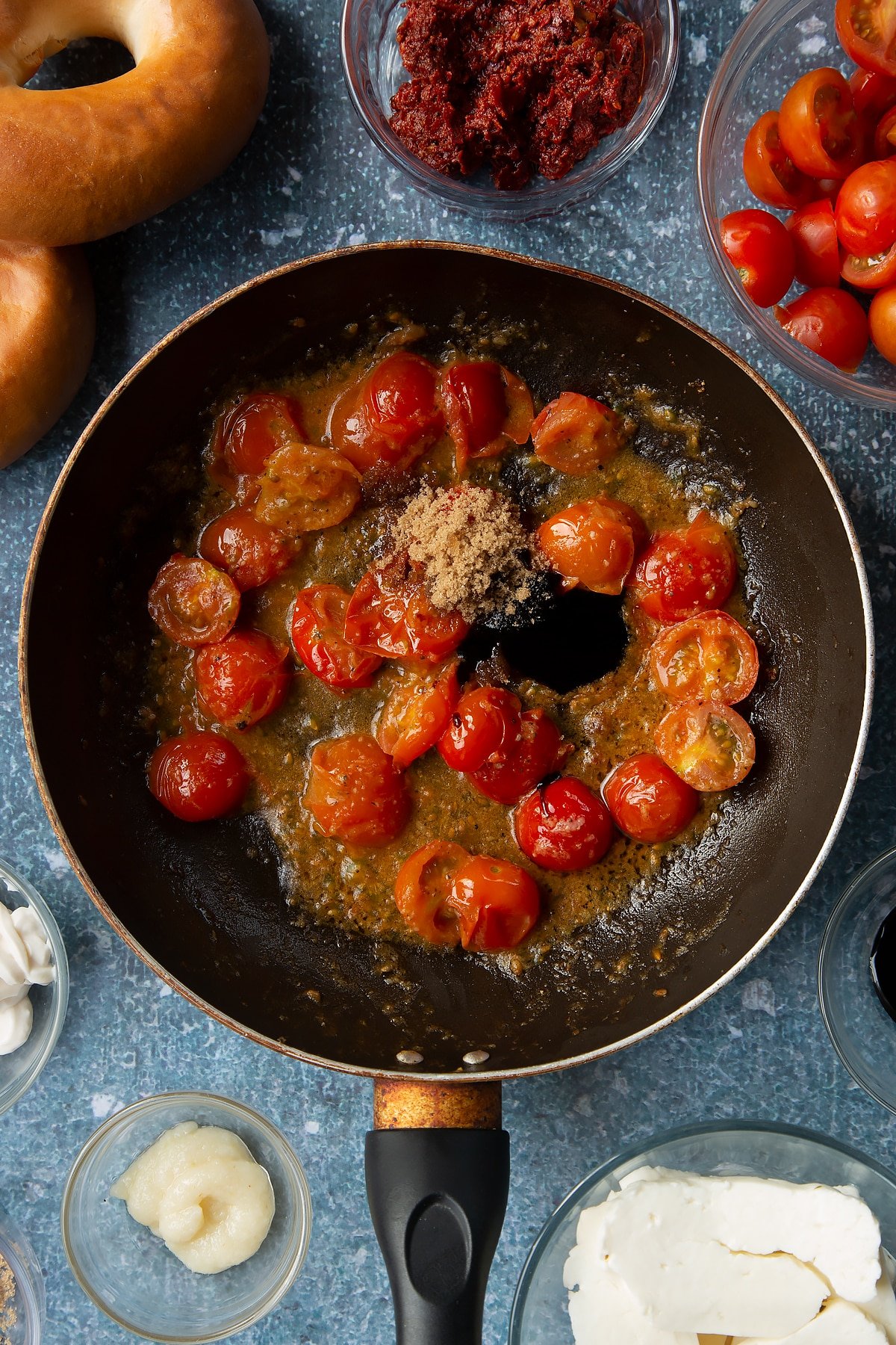 Fried down cherry tomatoes, garlic and pepper in a frying pan with brown sugar and balsamic vinegar added to it. Ingredients to make halloumi bagels surround the pan.