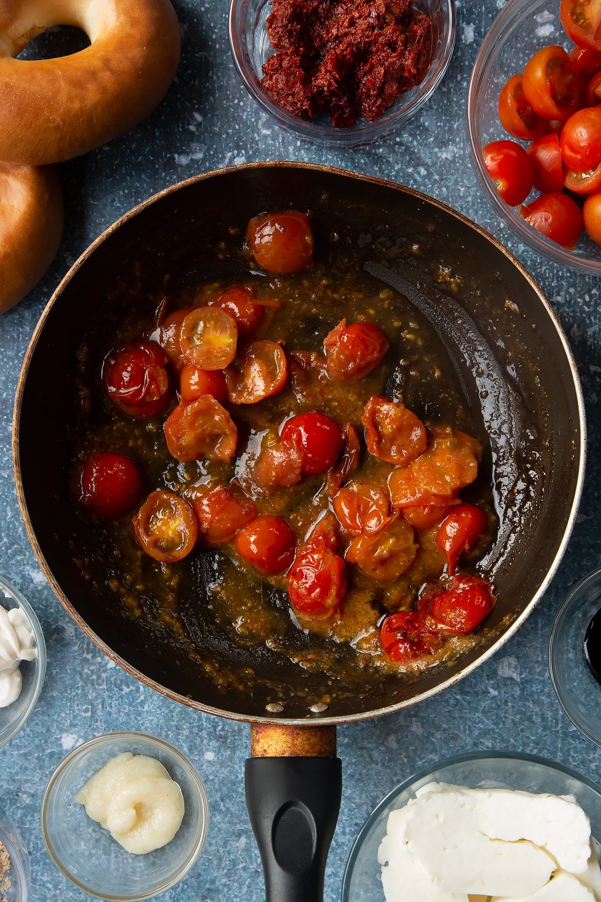 Fried cherry tomatoes, garlic, pepper, brown sugar and balsamic glaze in a frying pan. Ingredients to make halloumi bagels surround the pan.