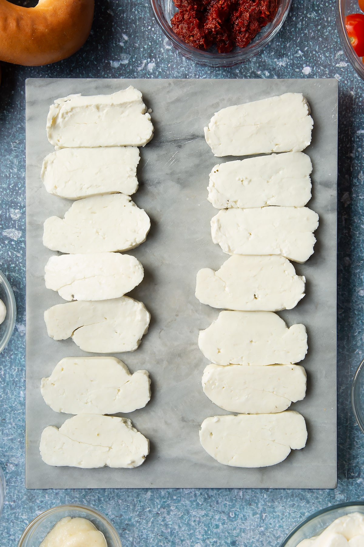 Slices of halloumi on a marble board. Ingredients to make halloumi bagels surround the board.