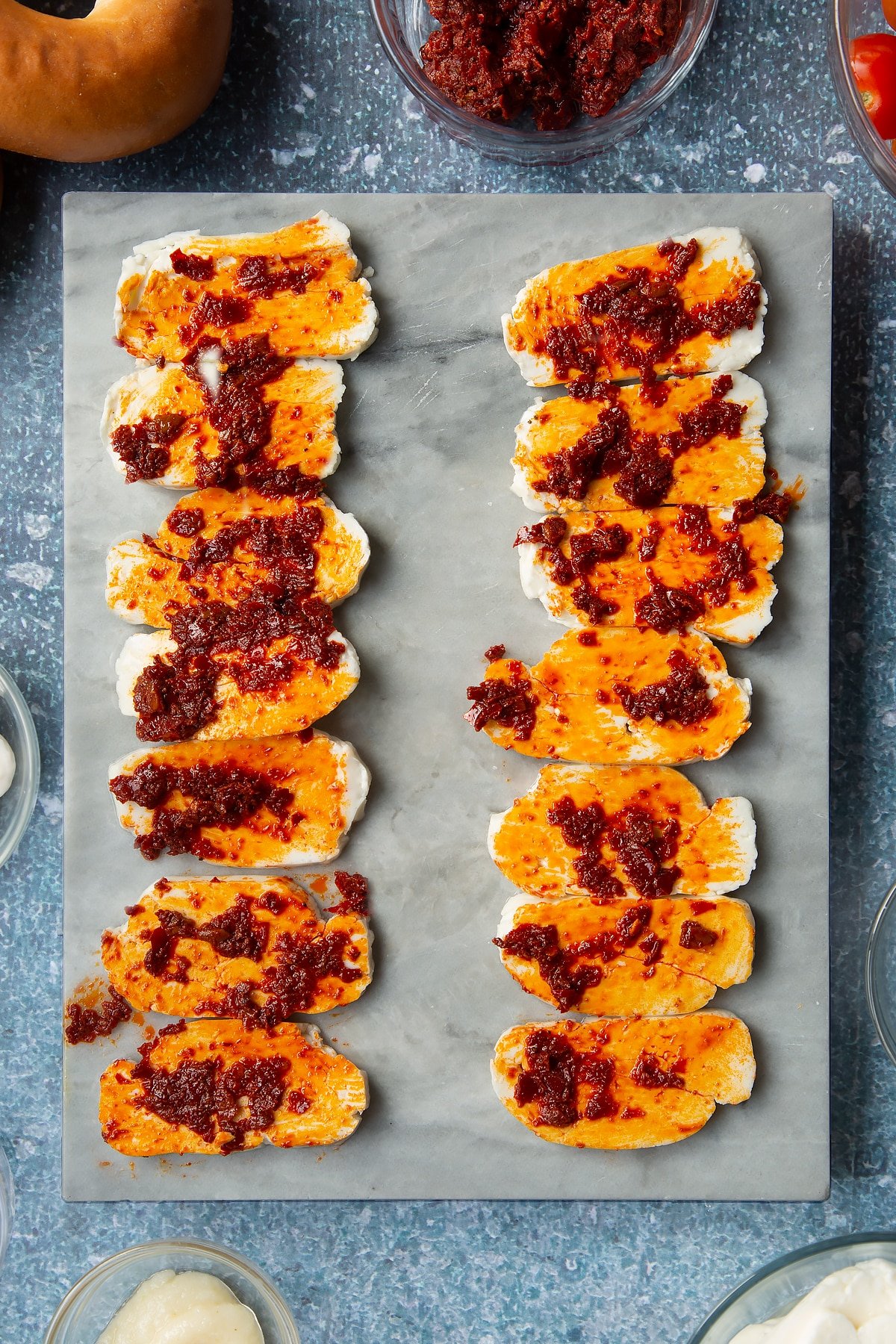 Slices of halloumi brushed with harissa paste on a marble board. Ingredients to make halloumi bagels surround the board.