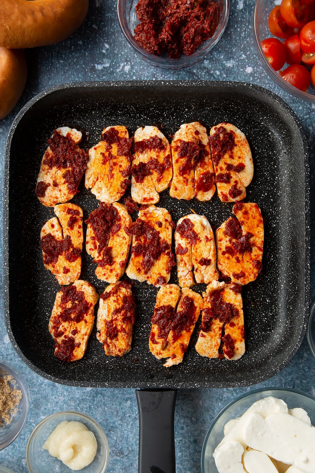 Slices of halloumi brushed with harissa paste in a griddle pan. Ingredients to make halloumi bagels surround the pan.