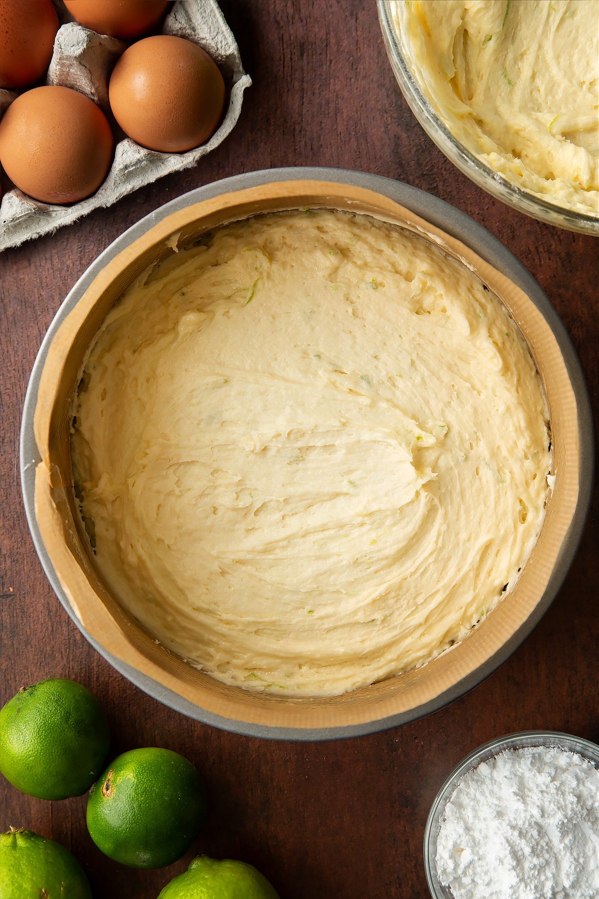 Lime drizzle cake batter in a lined cake tin. Ingredients to make lime drizzle cake surround the tin.