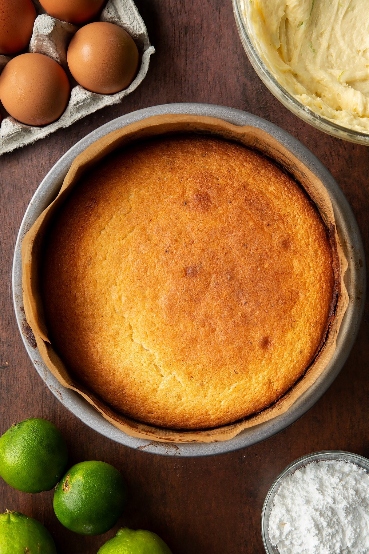 Baked lime drizzle cake in a lined cake tin. Ingredients to make lime drizzle cake surround the tin.