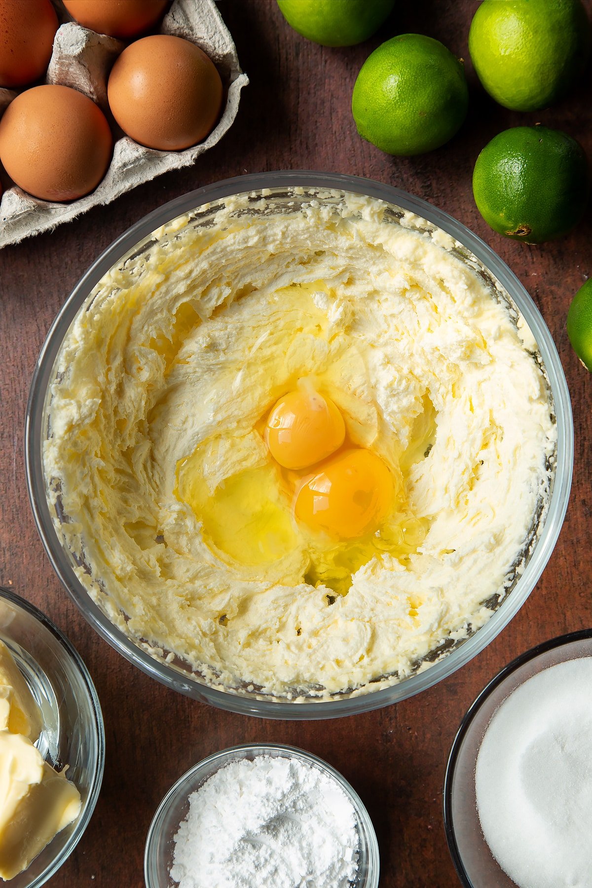 Butter and sugar beaten together in a mixing bowl with eggs on top. Ingredients to make lime drizzle cake surround the bowl.