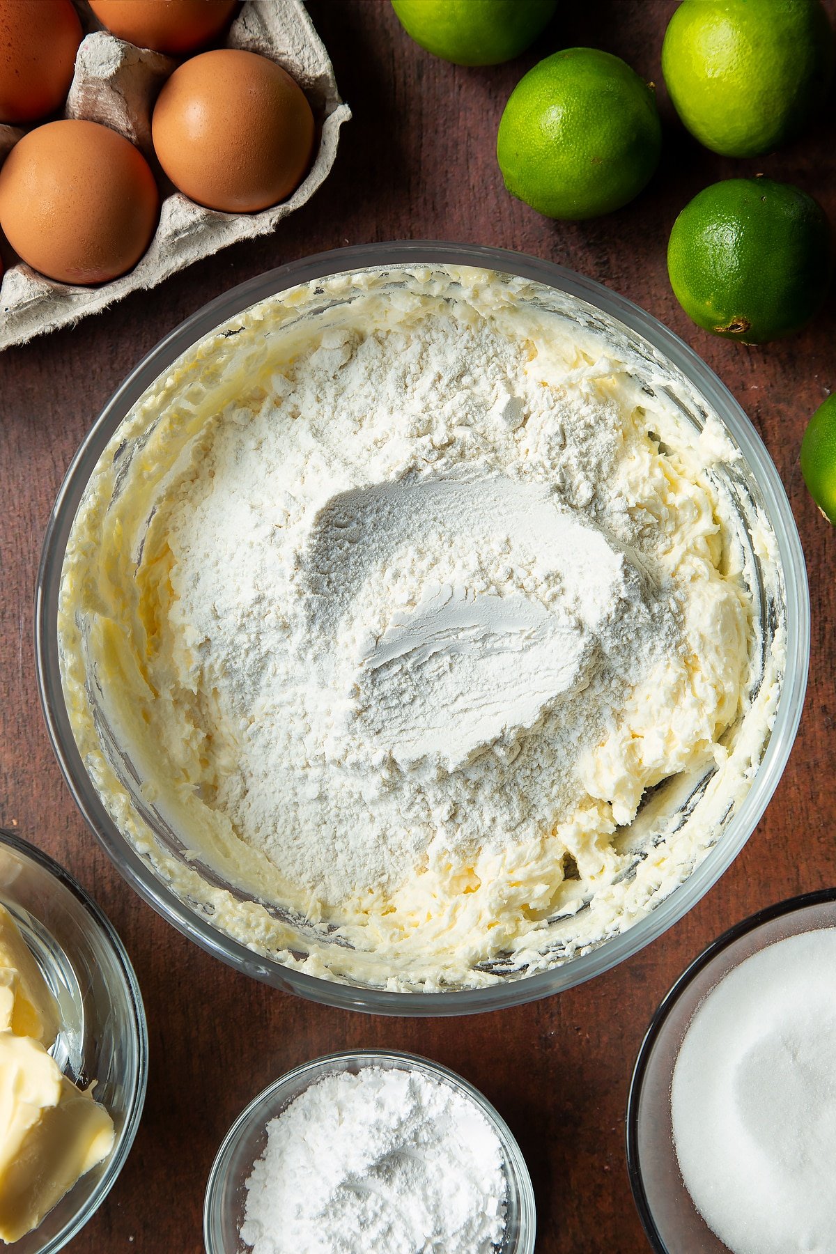 Butter, sugar and eggs beaten together in a mixing bowl with self-raising flour on top. Ingredients to make lime drizzle cake surround the bowl.