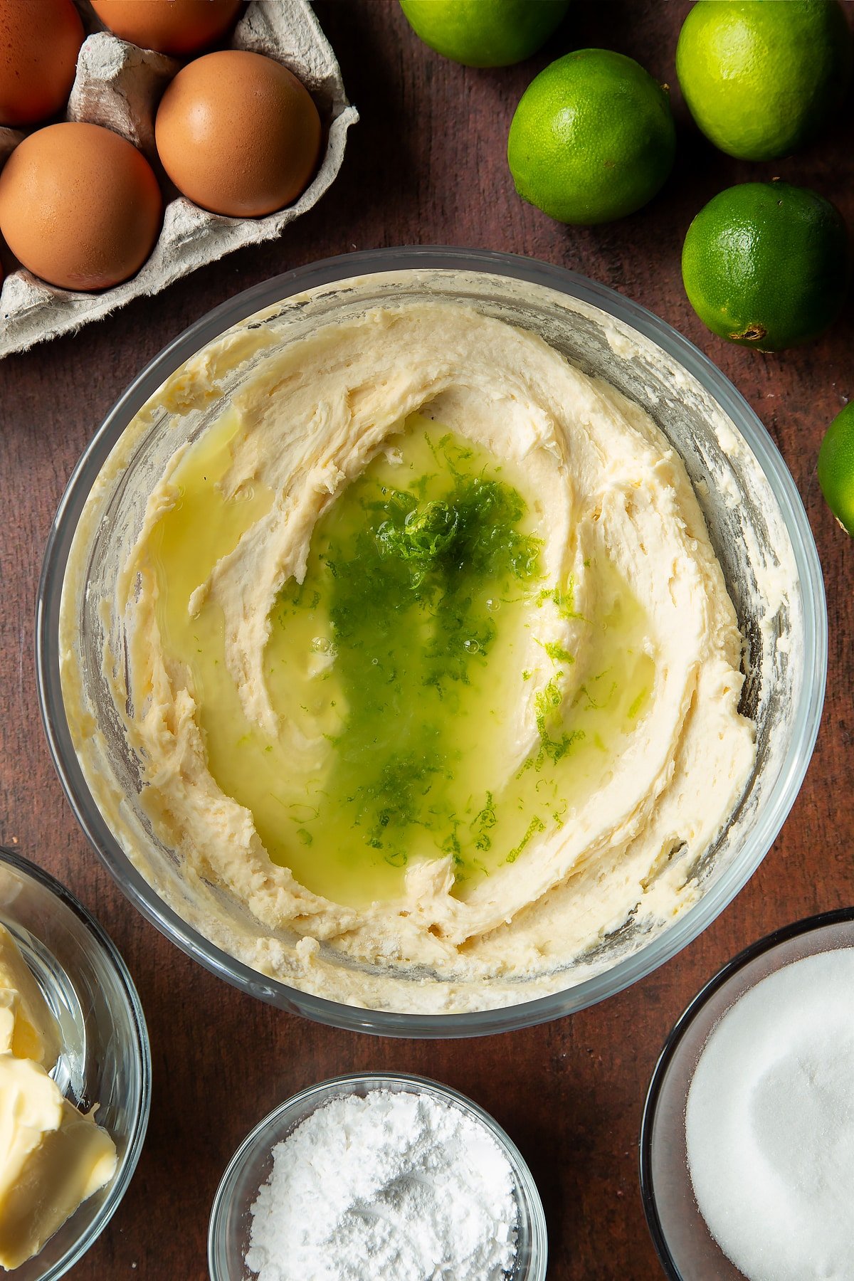 Basic cake batter in a mixing bowl with lime zest and juice on top. Ingredients to make lime drizzle cake surround the bowl.