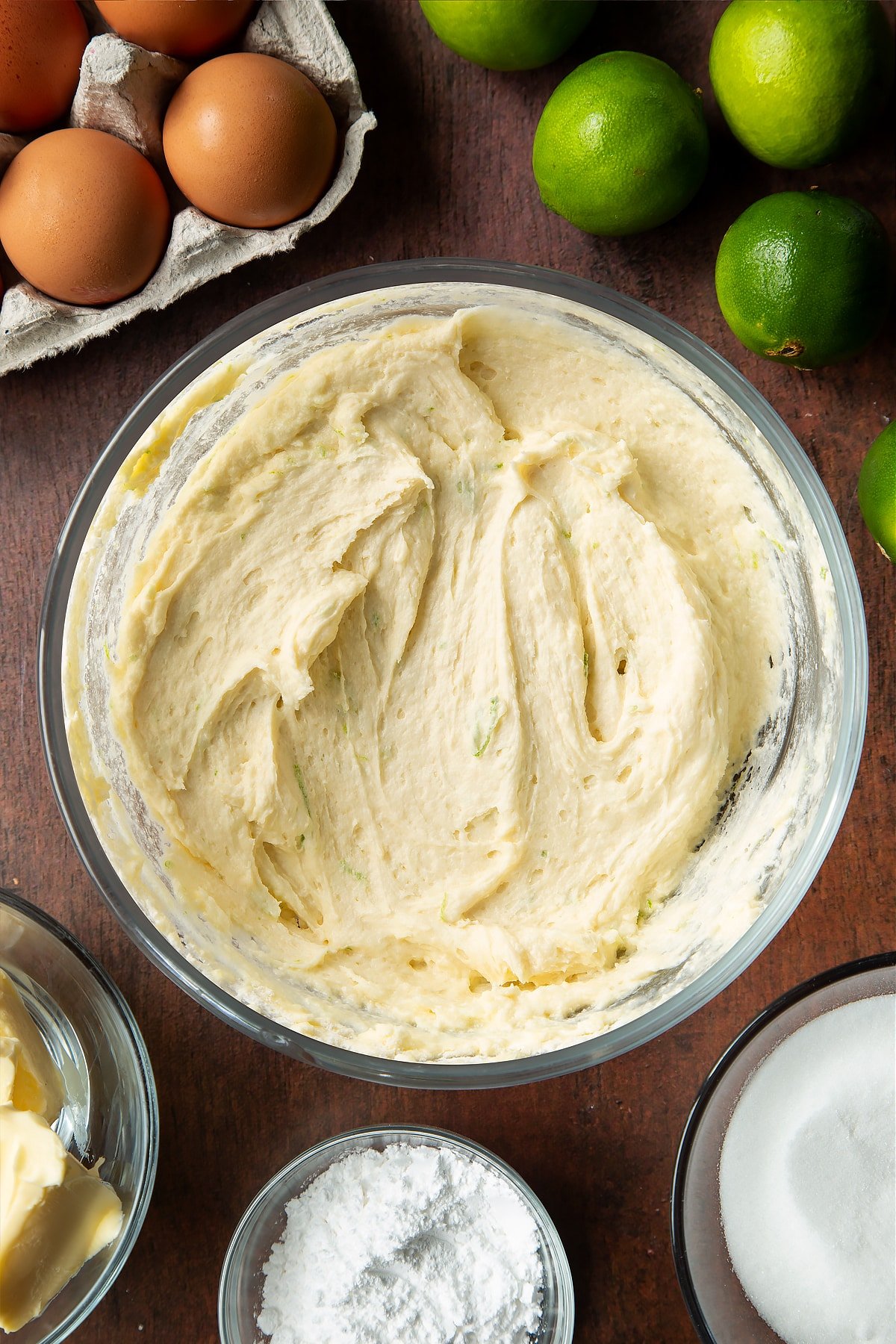 Lime drizzle cake batter in a mixing bowl. Ingredients to make lime drizzle cake surround the bowl.