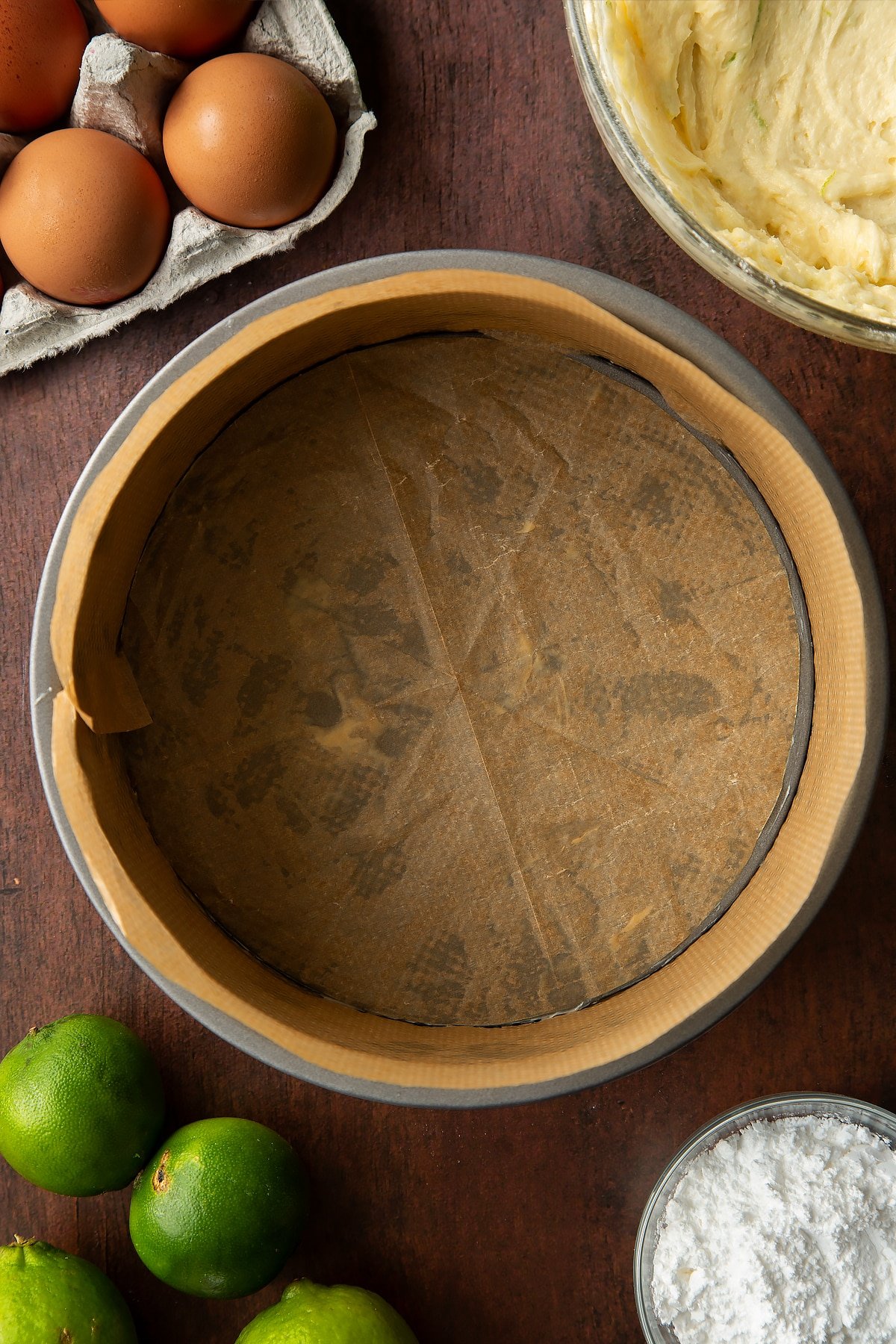 Cake tin lined with brown baking paper. Ingredients to make lime drizzle cake surround the tin.