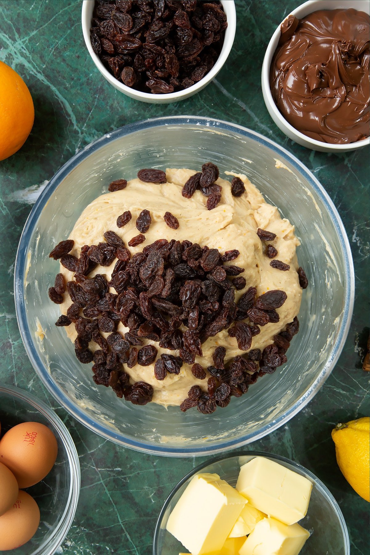 Spiced bread dough in a bowl with raisins on top. Ingredients to make Nutella panettone surround the bowl.