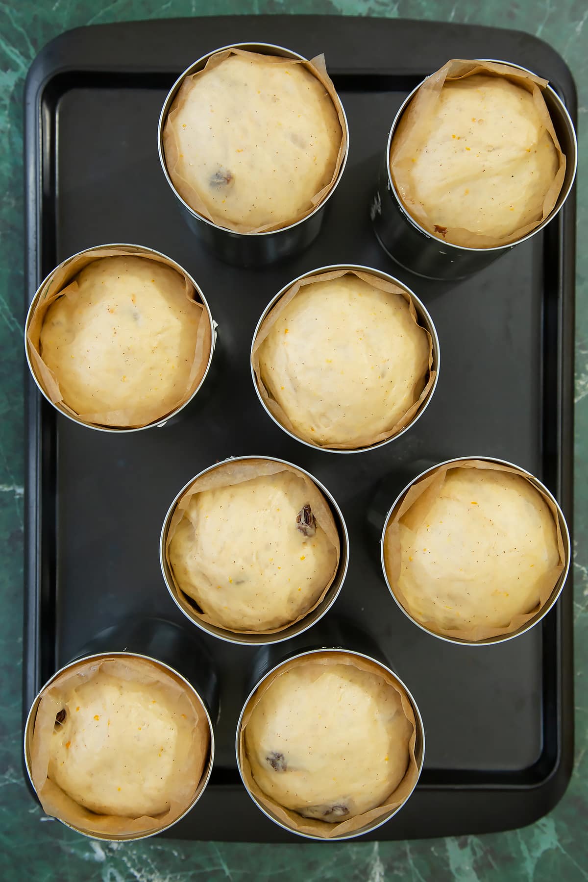 8 lined tin cans on a baking tray, each containing proved dough. 