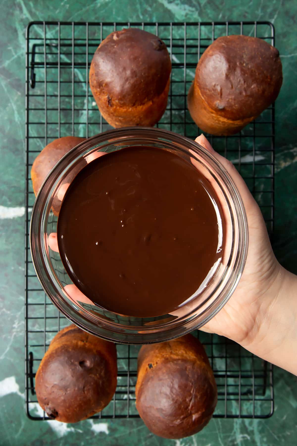 Hand holding a bowl containing melted dark chocolate. Below, 8 cooled panettone sit on a wire rack.