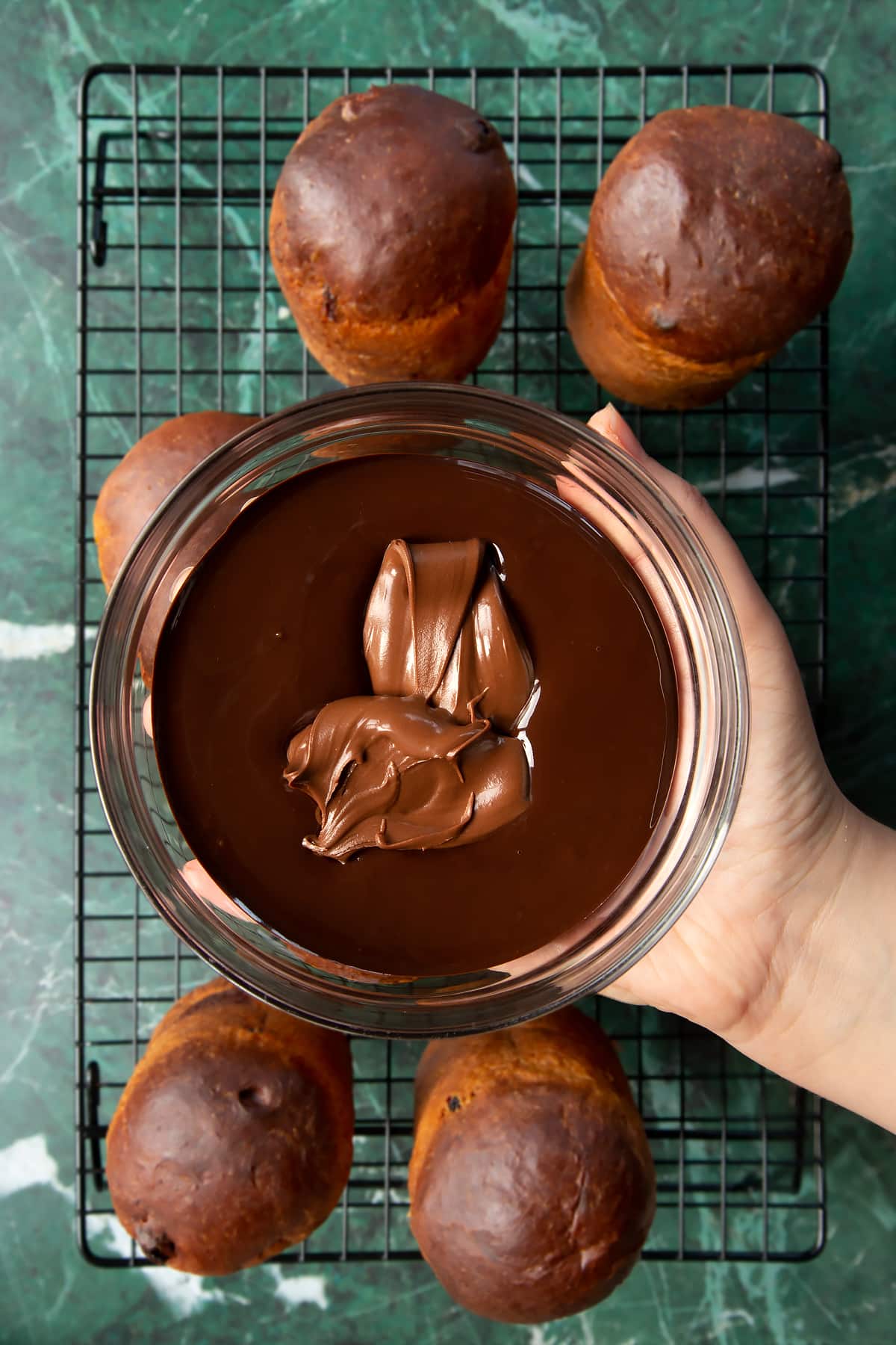 Hand holding a bowl containing melted dark chocolate and a scoop of Nutella. Below, 8 cooled panettone sit on a wire rack.