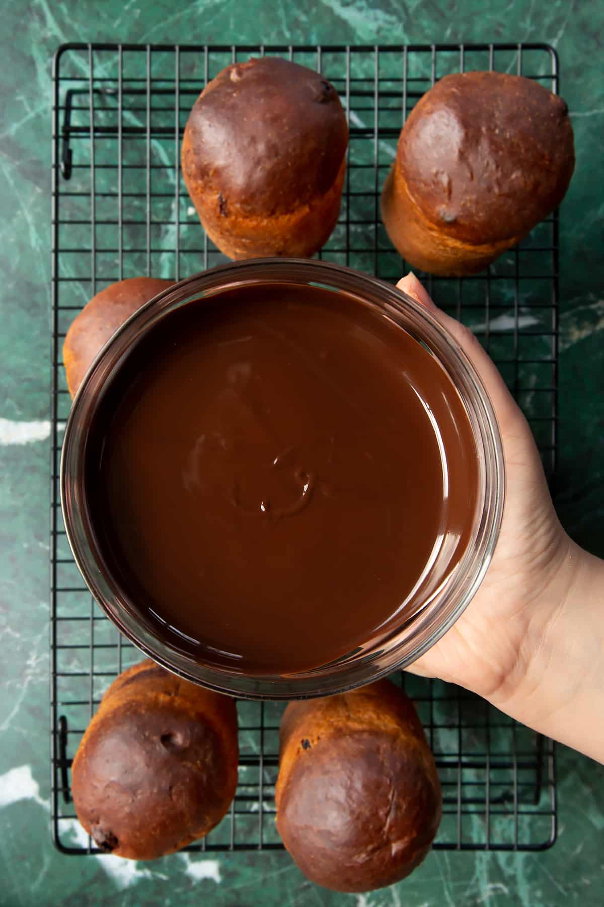 Hand holding a bowl containing melted dark chocolate and Nutella stirred together. Below, 8 cooled panettone sit on a wire rack.