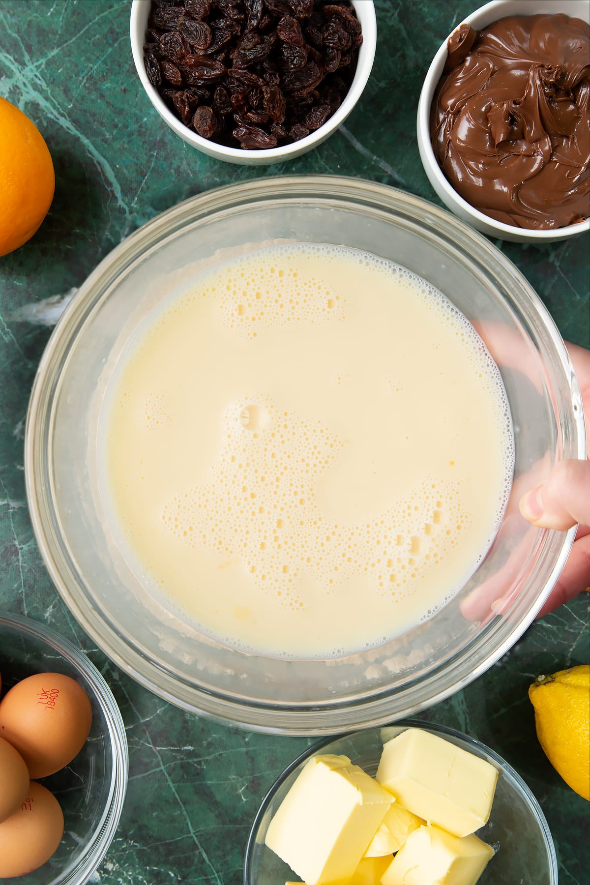 Hand holding a bowl of warm milk, eggs and vanilla whisked together. Ingredients to make Nutella panettone surround the bowl.