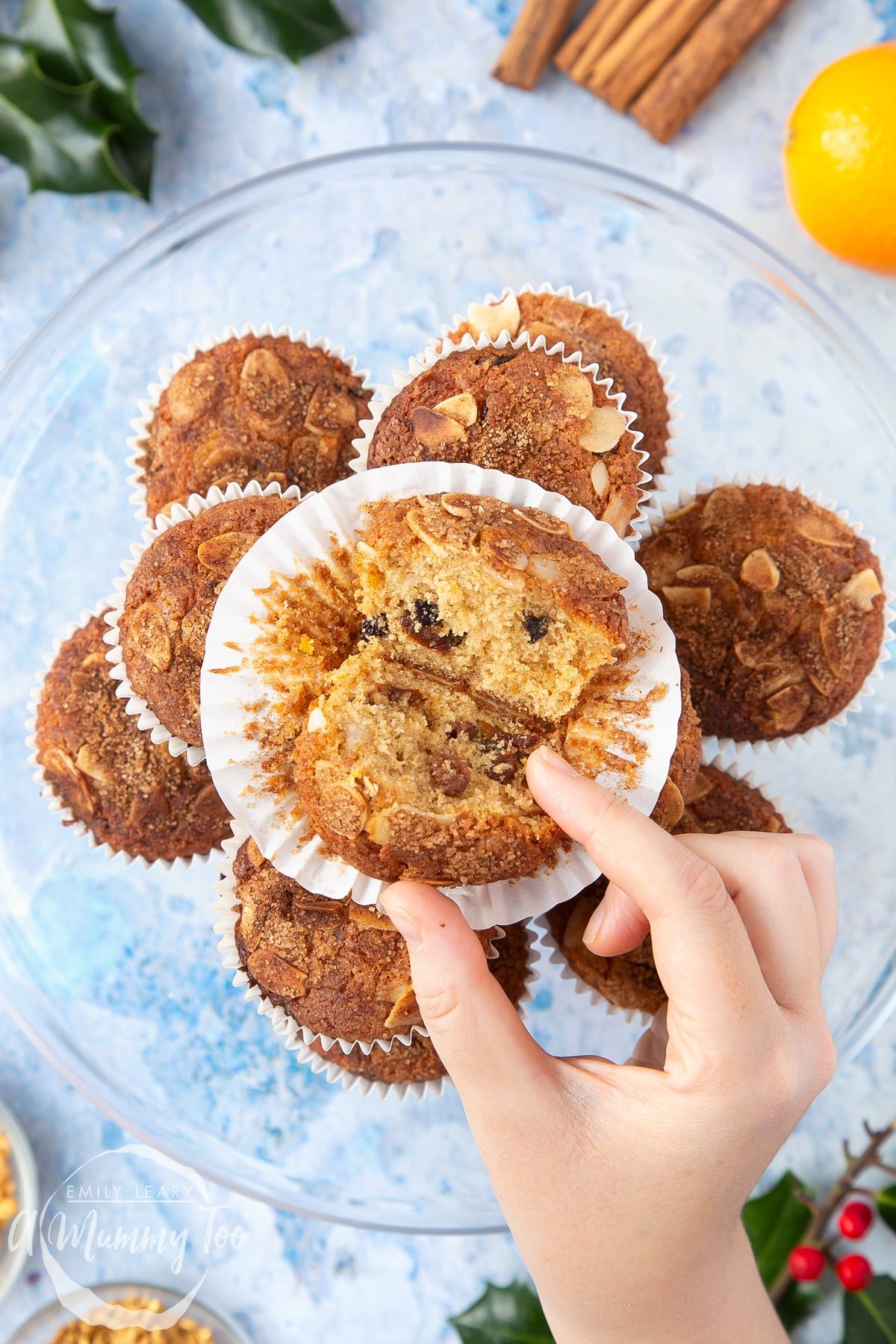 Overhead shot of a hand touching an uncased Orange and cinnamon muffin with a mummy too logo in the lower-left corner