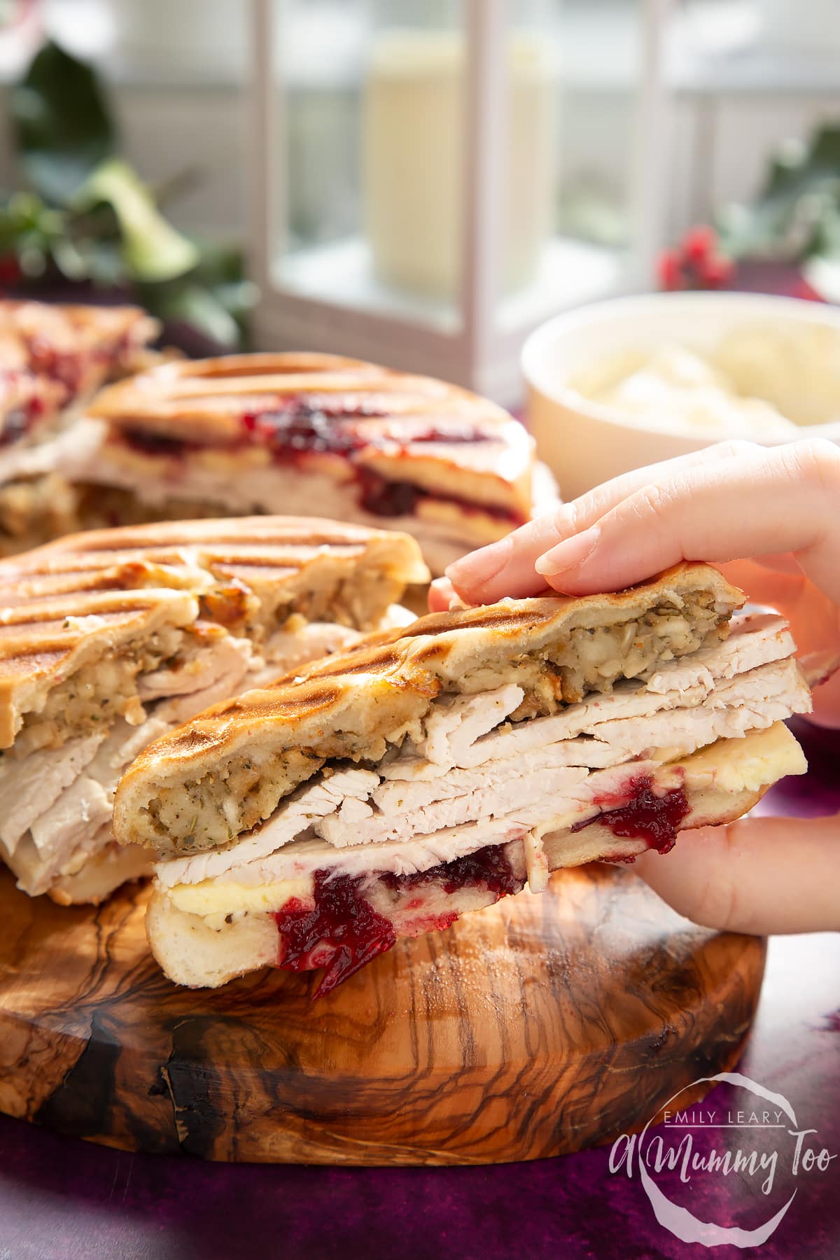 Front angle shot of a hand holding a Toasted turkey bagel served on a wooden plate with a mummy too logo in the lower-right corner